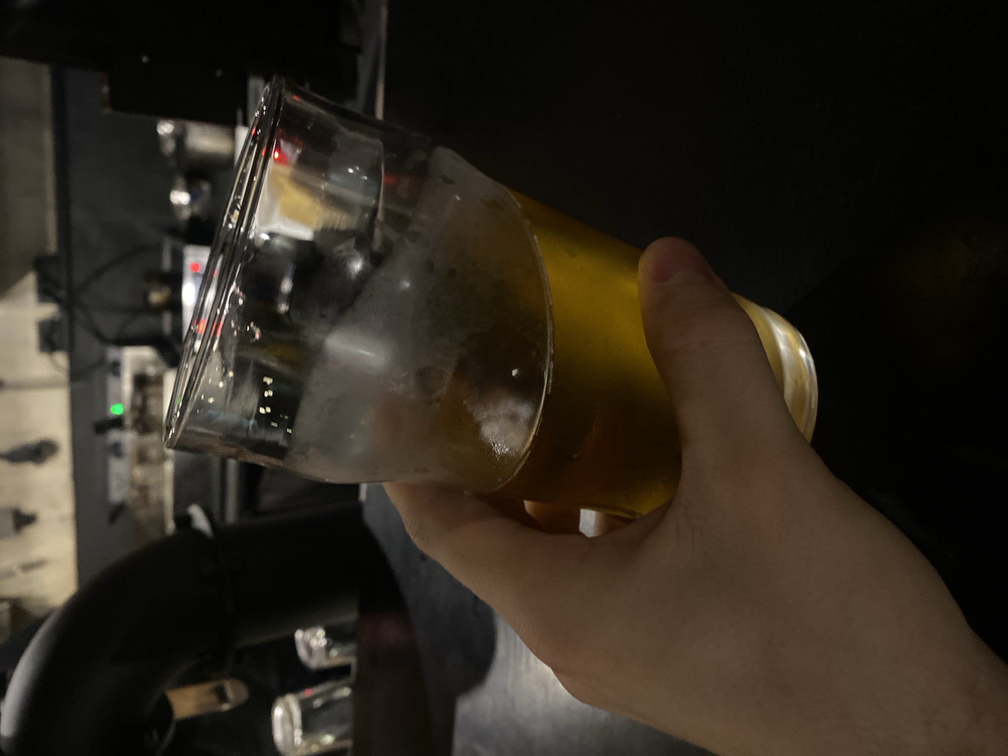 A hand holds a tilted glass of beer in a dimly lit environment. The glass is partially filled with beer, with a foamy head evident. The background reveals elements of a bar, including taps and possibly a draft beer system. The location in the city of Rusanovo, Russia is not apparent from the image.