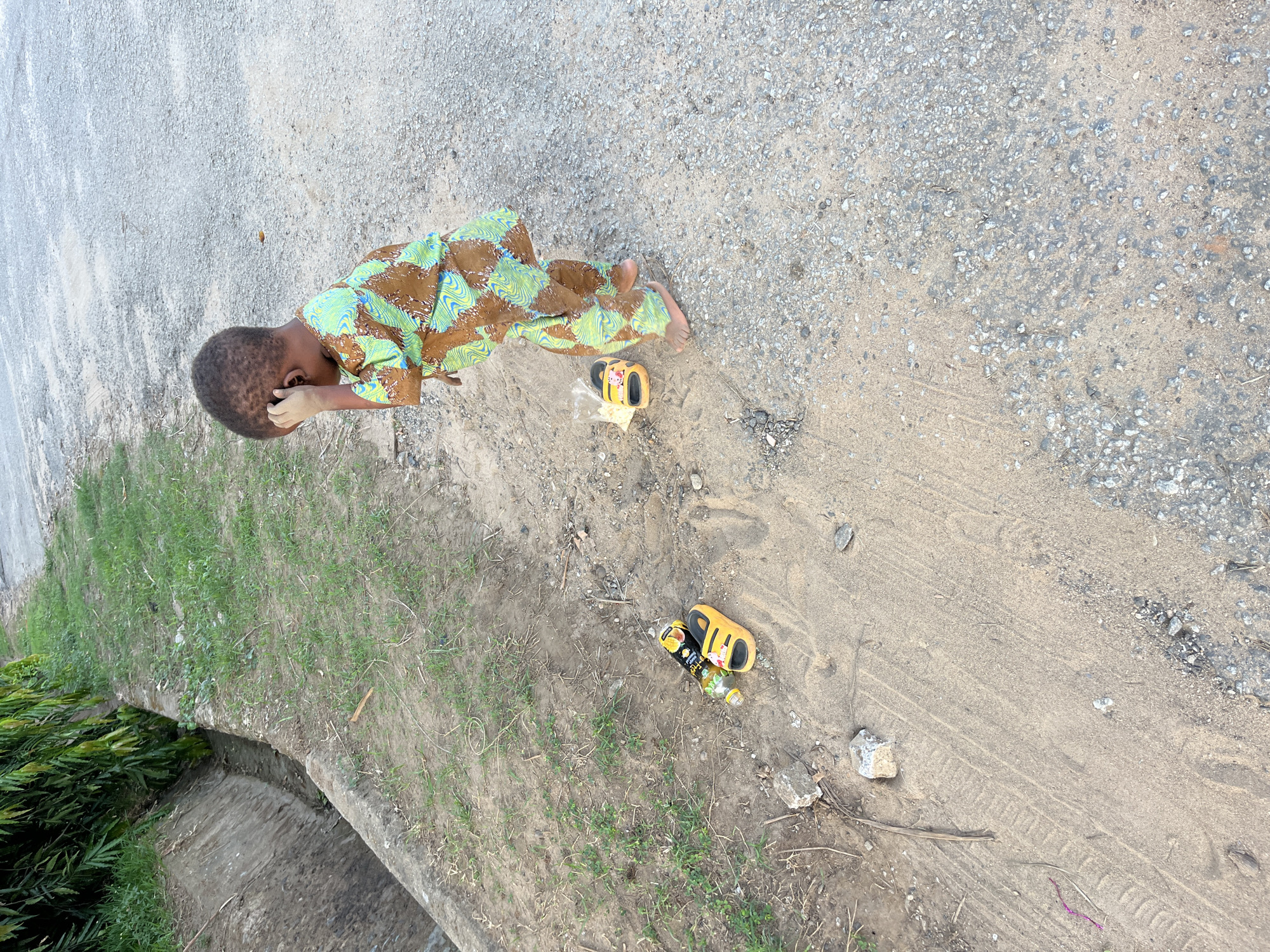 A young child, likely male, is lying on a partially paved and partially sandy ground in an outdoor location. The child is wearing a brown, green, and blue patterned long-sleeved shirt and pants. The child's left hand is touching the side of the head, near the ear.
Nearby are two pairs of children's yellow sandals. In proximity to one pair of sandals are plastic bottles. The ground is a mix of paved surface with gravel and a sandy area. There is green grass growing along the side of the paved area.
Based on the surroundings, it is probable the location is in a part of Abuja, Nigeria that is suburban.
