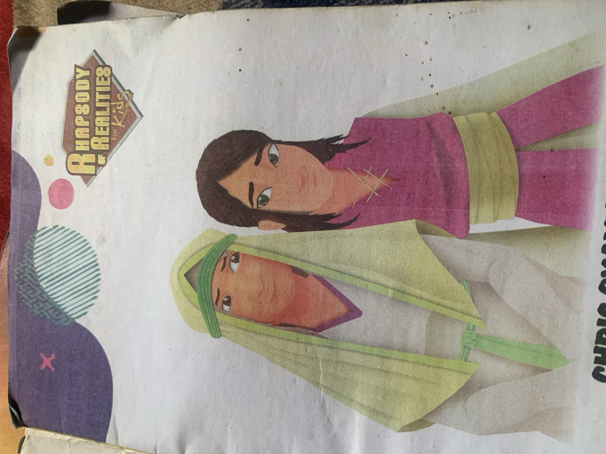 The image is a close-up, top-down photograph of a book cover. The cover features an illustration of two cartoonish figures, appearing to be of Middle Eastern or North African descent, standing side-by-side. The figure on the left wears a light green head covering and a white tunic with a purple trim. The figure on the right has dark hair and wears a pink tunic with a yellow sash. Both figures have warm skin tones and are depicted with expressive eyes.
At the top right of the cover, there is a logo that reads "RHAPSODY OF REALITIES for Kids" in a stylized font within a diamond shape. The background of the cover is a light, textured paper with some faint purple and circular green graphic elements at the top left. There is also a small pink "x" symbol in the top left corner of the cover. The bottom edge of the book cover is visible, with black text that appears to be a name, though it is mostly cropped out. The overall image quality is good, with clear details of the illustration.
There is no identifiable information in the image that would allow for its location to be determined within the city of Jauro Yinu, Nigeria, or any other specific geographical location. The image exclusively shows a book cover with an illustration and text.
