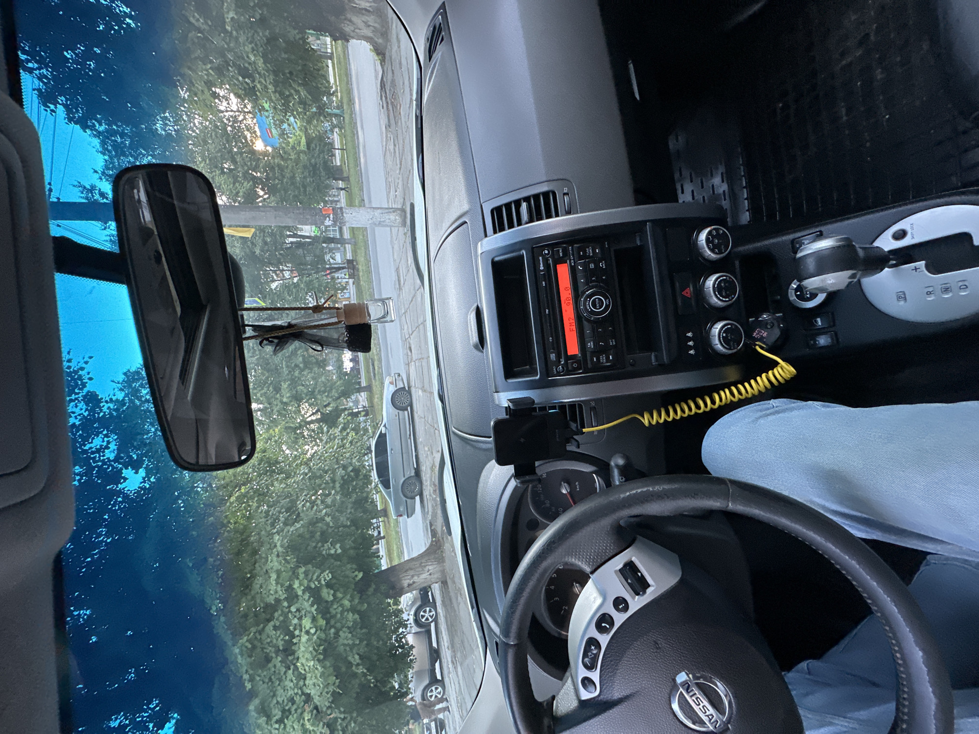 The image is a first-person view from inside a Nissan car, parked on a street in Kamianets-Podilskyi, Ukraine. The dashboard is visible, including the steering wheel with the Nissan logo, the radio display showing "FM2 90.0", the gear shift, and a yellow coiled cable. A person's legs in light blue jeans are visible on the driver's side. Outside the car, through the windshield and side windows, trees and other parked cars are visible. A grey sedan is parked on the right side of the road, and another vehicle, possibly an SUV, is parked on the left. The environment appears to be a city street with trees lining the sidewalk. The sun is setting, casting a warm light on the scene.