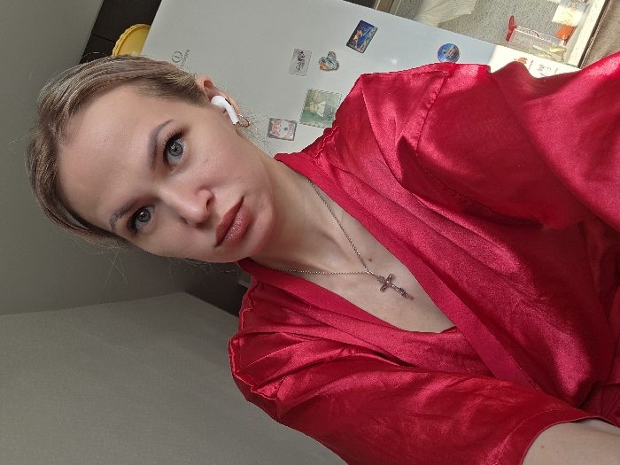 A female subject, adult, with light hair pulled back and light eyes, is depicted in a front-facing, chest-up shot. She wears a bright red satin robe and a silver chain necklace with a crucifix pendant. A white wireless earbud is present in her right ear, along with a small gold hoop earring. Her gaze is directed at the camera, maintaining a neutral facial expression with glossy lips. The scene is an indoor domestic environment, identifiable as part of a kitchen or utility area within the city of Chaikovsky, Russia. To the subject's right, a white Indesit refrigerator is visible, adorned with numerous magnets of various designs, and a yellow container rests on its top. The background to the left consists of a plain light-colored wall. Additional small objects are discernible on a surface in the lower right portion of the image.