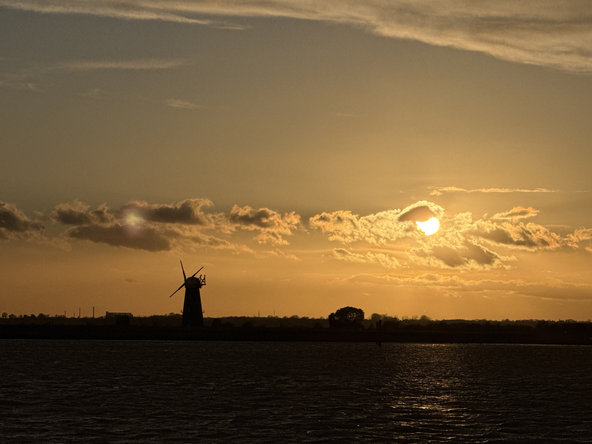 The image displays a panoramic landscape at sunset or sunrise over a body of water in the vicinity of Great Yarmouth, United Kingdom, likely along the River Yare or within the Norfolk Broads. The upper half of the frame is filled with a sky rendered in shades of golden-orange, transitioning to a lighter hue at the zenith. The sun, a bright, partially obscured orb, is positioned in the upper right quadrant, veiled by layered clouds that exhibit strong backlighting and golden illumination. Thin, linear clouds are visible in the upper sky.
The lower half of the image comprises a dark, rippled expanse of water, reflecting a vertical column of shimmering sunlight that extends from the horizon towards the foreground. A distinct, horizontal landmass appears as a dark silhouette across the mid-ground. On the left side of this landmass, a four-sailed windmill, consistent with traditional Norfolk mills such as Berney Arms Windmill, stands prominently as a dark silhouette. To the right of the windmill, along the same horizon line, a dark tree line is visible, interspersed with several faint, distant industrial structures, including vertical stacks. There are no human figures, animals, or discernible interactions present in the scene. The environment is static and appears to be a rural or semi-rural waterway.