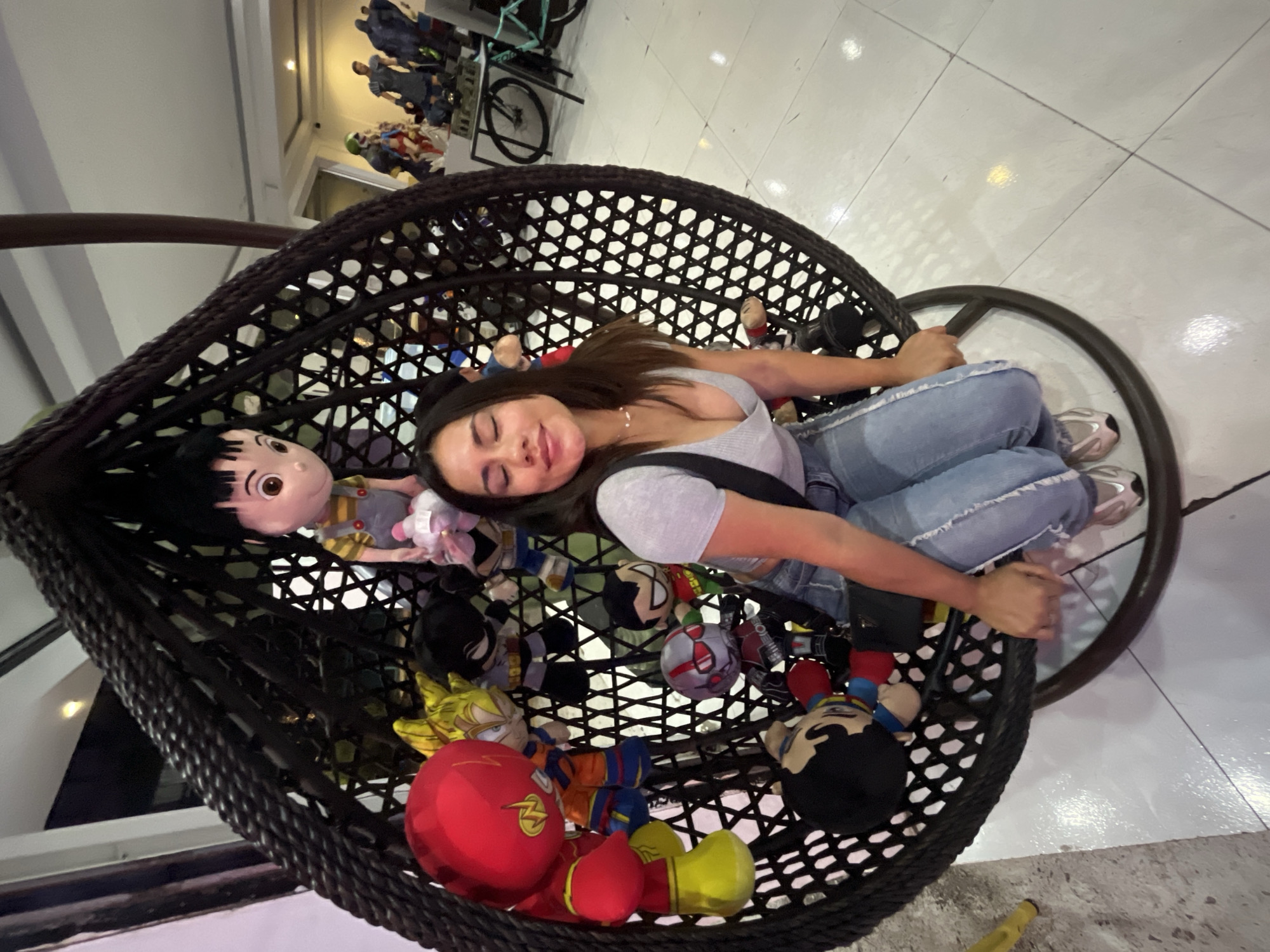 A high-angle shot reveals a woman reclining in a hanging wicker chair. She is fair-skinned with long, dark hair, wearing a light-grey top and light-blue jeans, and silver sneakers. The chair is densely filled with a variety of cartoon character plush toys, including what appears to be a Flash doll.
The chair's frame is dark and crafted from wicker. The floor is tiled in white. The background shows partial views of toys displayed on shelves and a bicycle hanging from the ceiling, suggesting an indoor environment, potentially a retail space or a private residence. The location is Unknown, Unknown.