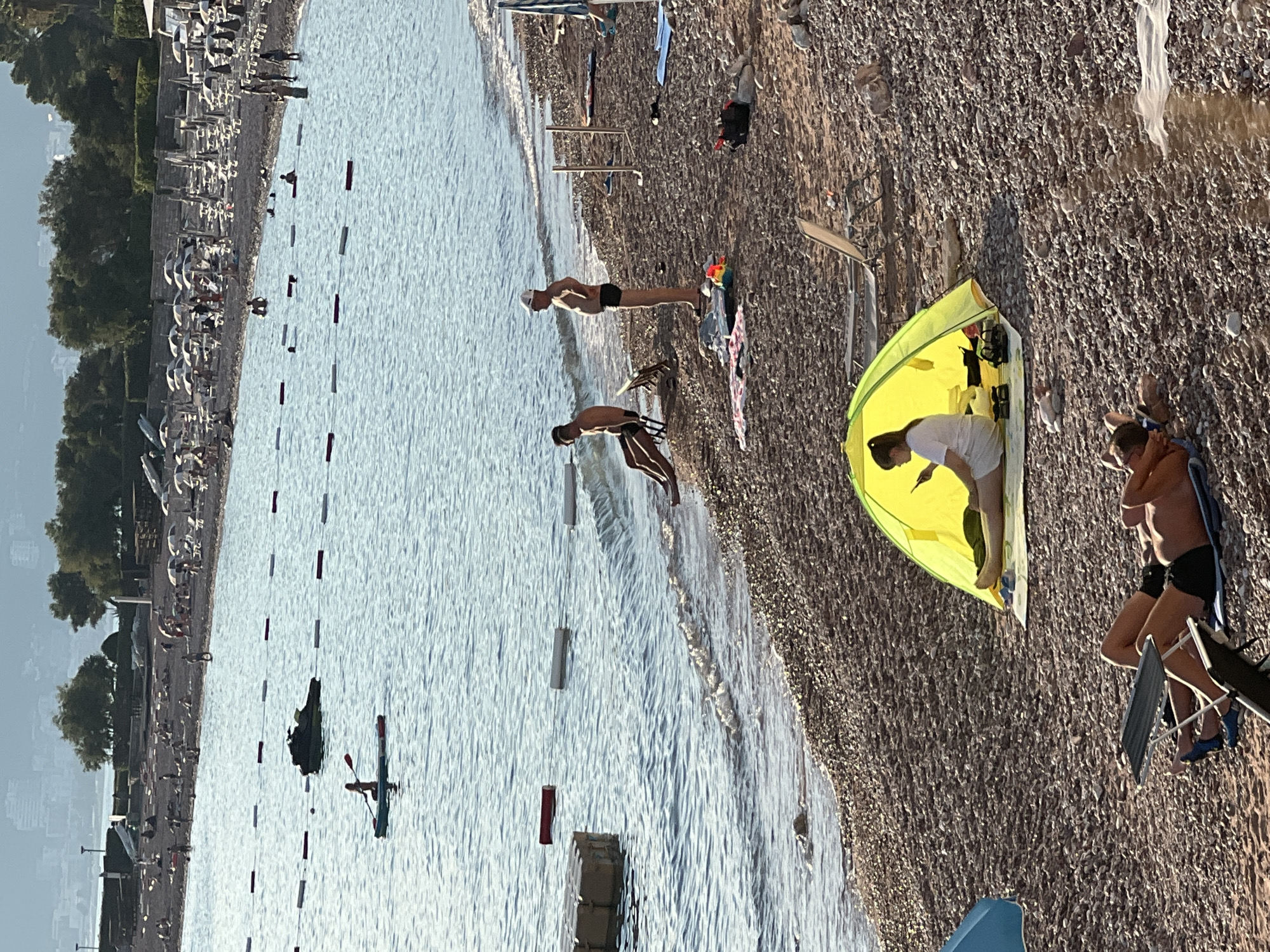 In Sveti Stefan, Montenegro, a crowded pebble beach lines a calm, blue sea. Several people are visible on the beach and in the water. In the foreground, a person reclines on a lounge chair under a blue umbrella. To the right, a bright yellow tent shelters a person using a mobile phone. Further back, two men are seated in chairs near the water's edge, while another man stands nearby. The water has a floating barrier marked with white and red buoys, and in the distance, a jet ski is parked. The background shows a tree-lined shoreline with buildings and numerous beach umbrellas and lounge chairs.