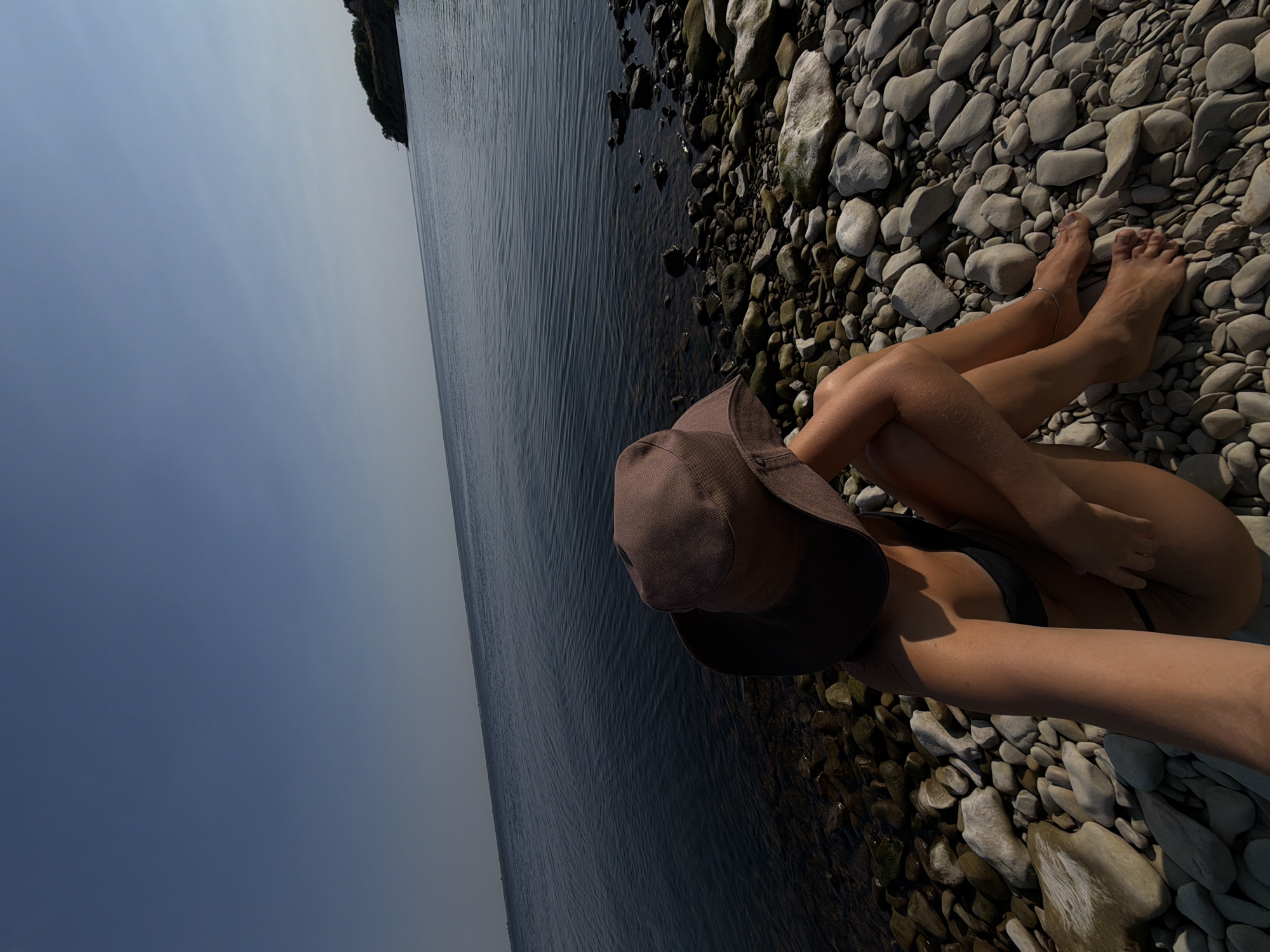 A woman wearing a dark-colored bikini and a wide-brimmed hat sits on a rocky shoreline, facing a dark, calm body of water. Her head is tilted down, and her left arm is resting on her knee. The shore is covered in smooth, grey and white stones of various sizes. In the distance, the water meets a clear blue sky, with a small, green, tree-covered island or peninsula visible on the right horizon. The image appears to be taken during daylight. The location is not identifiable as being in the city of Kamyshin, Russia, based on the visual information present.
