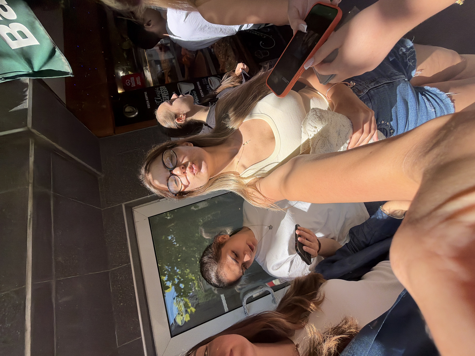 The image shows a group of young women in an outdoor setting, possibly in Burgas, Bulgaria. One woman in the foreground is taking a selfie, puckering her lips and wearing glasses and a light-colored tank top. She is holding a white lace bag. To her left, another woman with long brown hair is visible from the chest up, wearing a white top and blue jeans. Behind them, a woman with dark hair pulled back is looking down at her phone, wearing a white t-shirt. Further back, a man in a white t-shirt is visible. The background suggests a commercial area with a building facade, a window with reflections of trees, and signage that appears to be for "Costa Coffee." A green awning with the letter "B" is visible at the top right. The lighting indicates it is daytime, with strong sunlight casting shadows. One woman is holding an orange-cased smartphone.
