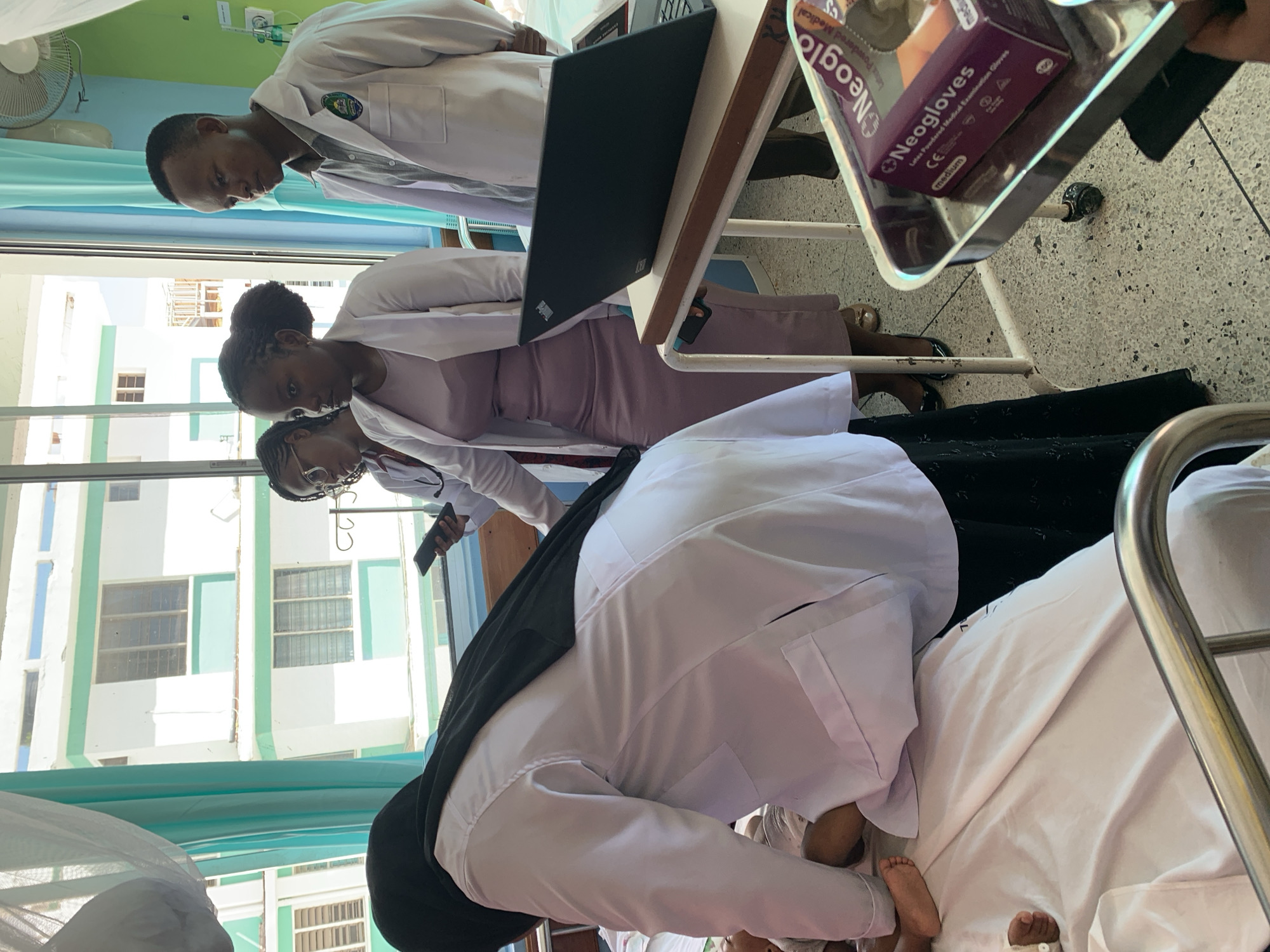 **Location:**
The scene appears to be taking place inside a hospital room. The presence of hospital beds, medical equipment, and uniformed medical personnel suggest a medical setting, likely in Dar es-Salaam, Tanzania.
**People:**
The image shows multiple individuals, primarily medical personnel. They are dressed in white coats. The presence of medical students or trainees cannot be ruled out.
**Subjects:**
A patient is in the hospital bed, presumably receiving medical attention.
**Objects:**
Objects present in the scene include a laptop, medical gloves ("Neogloves" brand), a medical tray, a fan, and standard hospital room fixtures (such as curtains, windows, and equipment).
**Actions and Interactions:**
Medical personnel are gathered around the patient's bed. They seem to be involved in a medical examination. Some are looking at a laptop, potentially reviewing patient data or diagnostic images. The woman in the black head covering is bent over the bed, appearing to attend to the patient.
**Environment:**
The environment is a typical hospital room setting. The room has windows that overlook an exterior building.