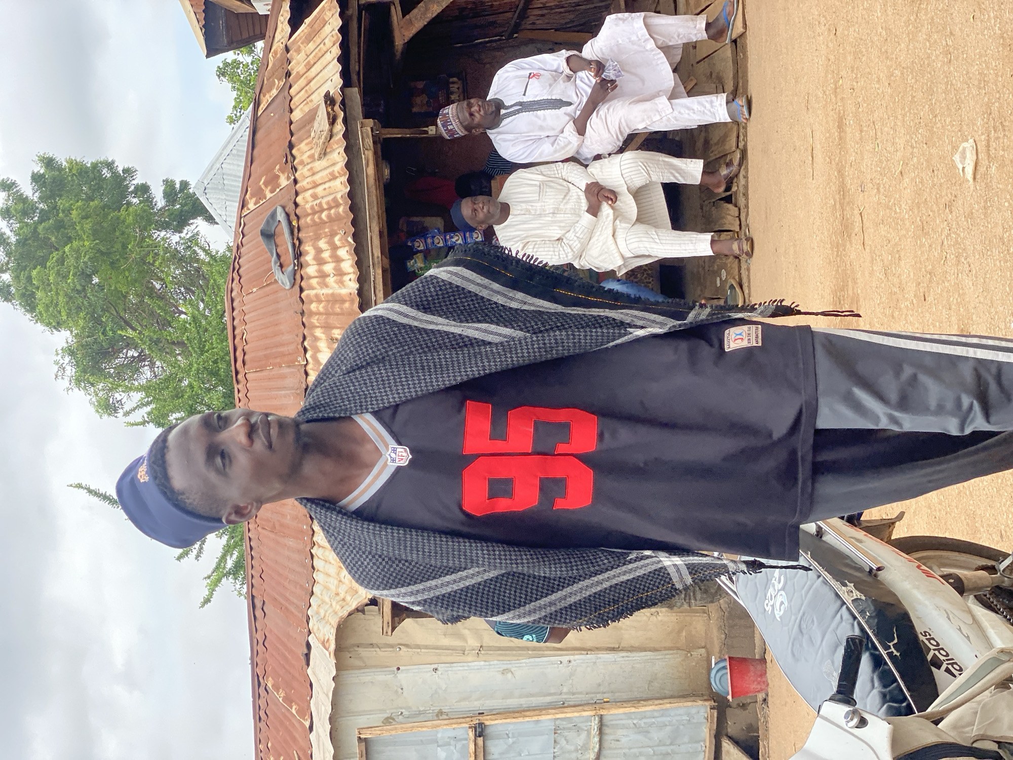 In Rigachikun, Nigeria, a young man wearing a blue cap, a black jersey with the number "95" in red, and a patterned shawl stands in the foreground, looking to his right. Behind him, a weathered, corrugated tin roof structure, likely a shop, is visible with two older men sitting on a wooden bench. One man wears a white striped kaftan and the other a white kaftan with intricate embroidery, both wearing traditional caps. A motorcycle is partially visible in the lower left corner of the frame, with a red bucket in the background. The ground is composed of reddish-brown earth.