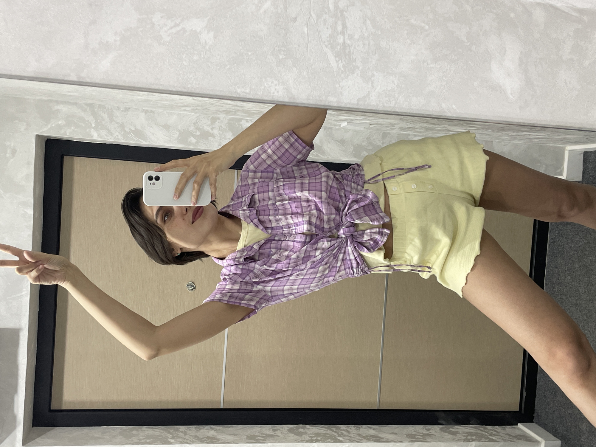 This image is a mirror selfie taken by a young woman in an indoor setting in Izhevsk, Russia. The woman, with dark, shoulder-length hair and fair skin, is positioned slightly off-center to the right. She holds a white smartphone in her right hand, aiming it towards the mirror to capture her reflection. Her left hand is raised, forming a peace sign, suggesting a casual and possibly playful mood. Her facial expression is neutral, with her lips slightly parted as she looks towards the phone.
She is wearing a casual outfit consisting of a purple and white plaid short-sleeved button-up shirt, which is tied in a knot at her waist. Beneath the plaid shirt, a light yellow or cream camisole or top is visible. She also wears light yellow or cream shorts that feature a subtly ruffled hem and a purple drawstring.
The background visible in the mirror's reflection consists of architectural elements. Behind the woman, a light brown or tan door with a prominent black frame is seen. To the left of the door, and covering the upper portion of the reflected space, is a light grey wall with a distinct textured finish, possibly a decorative plaster or wallpaper. The floor, visible in the lower part of the reflection, appears to be a dark grey, suggesting carpet or tile. The bright and even lighting indicates either daytime or a well-lit artificial environment. No visible text is present in the image.