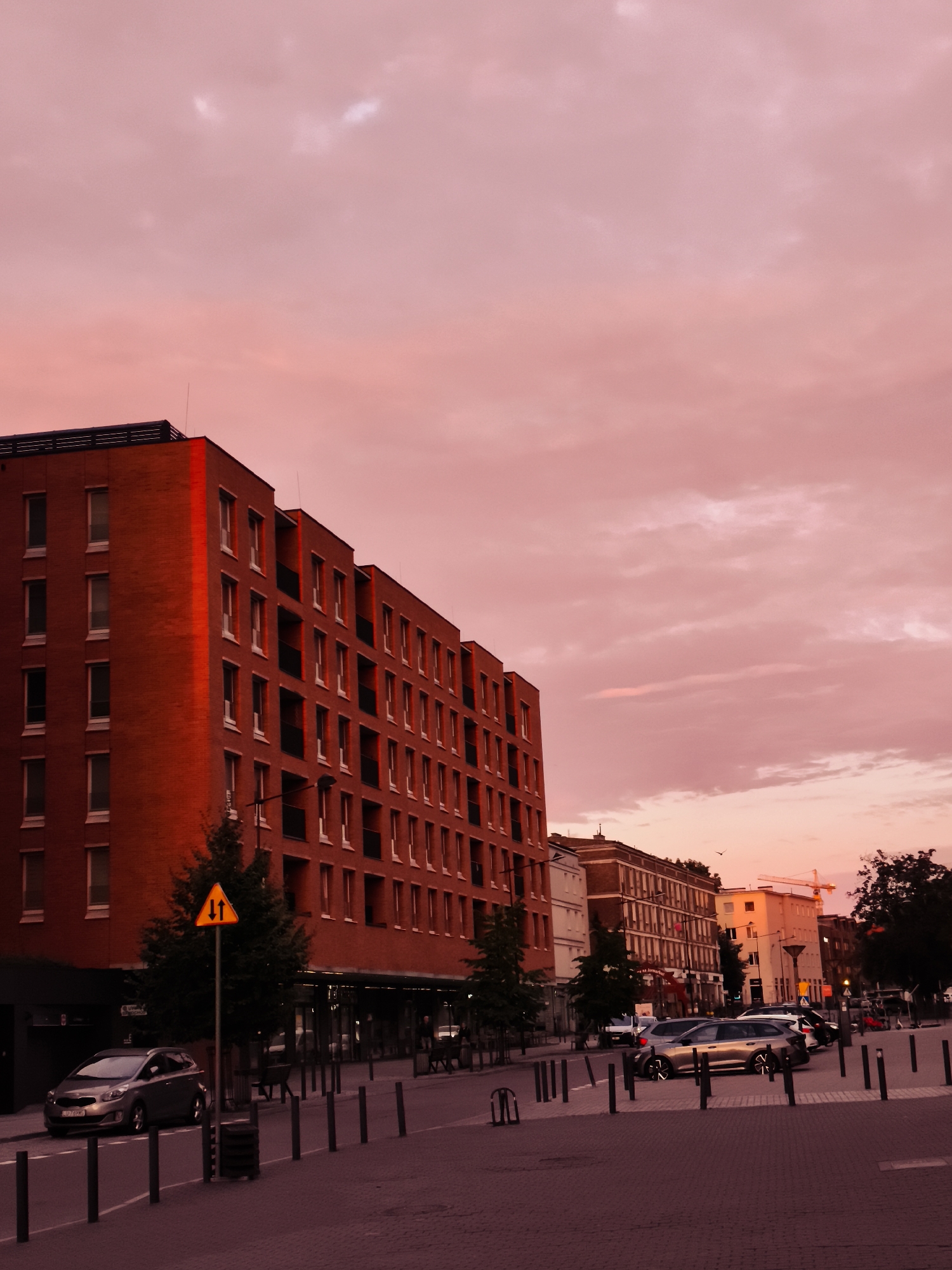 A brick apartment building with many windows and balconies dominates the left side of the frame. The building is bathed in the warm, reddish light of a sunset, which also colors the sky and clouds with shades of pink and orange. A silver hatchback car is parked on the street in front of the building. A traffic sign with two arrows indicating two-way traffic is placed on a pole near a small evergreen tree. In the background, other buildings are visible, including a taller building with a crane on top. The street is paved with brick, and there are bollards lining the sidewalk. The scene appears to be in an urban environment, possibly Gdańsk, Poland, given the context.