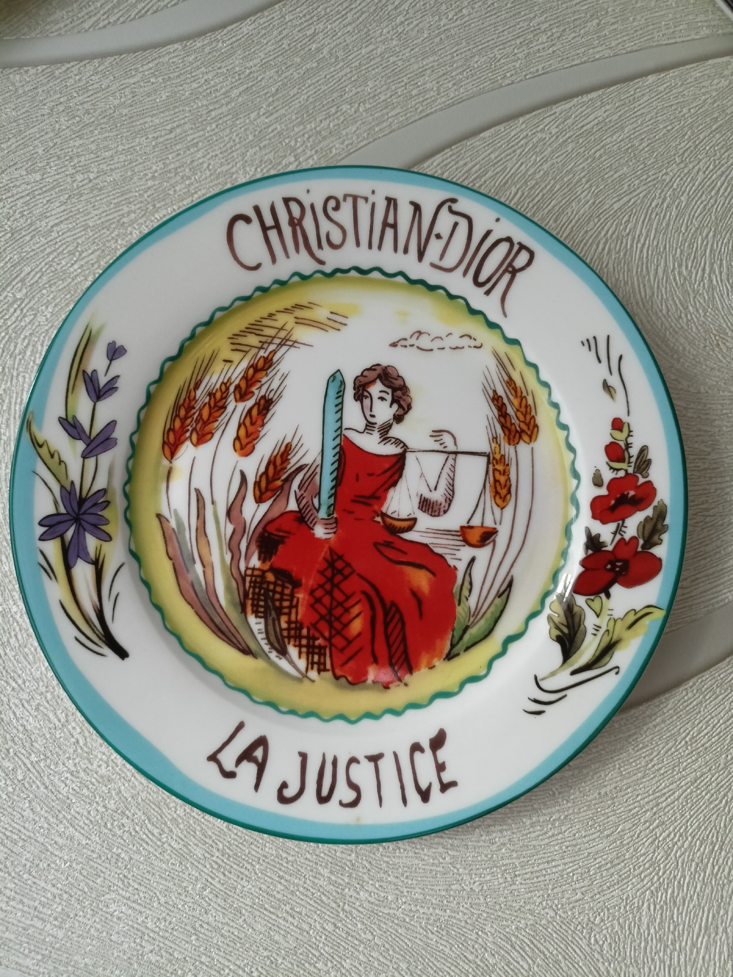 A ceramic plate with a decorative design is displayed on a textured surface, likely a tablecloth or placemat. The plate features a central illustration of a woman, presumably representing justice, holding scales in one hand and a blue rod in the other. She is dressed in a flowing red gown and is depicted amidst representations of wheat stalks. The words "CHRISTIAN DIOR" are inscribed in a curved line above the central image, and "LA JUSTICE" is written below it. The plate has a decorative border with floral motifs on the left and right sides, and a turquoise trim along its edge. The background of the image is a neutral, textured fabric. There are no people present in the image. The image is an overhead shot of the plate. The location in the city of Volgodonsk, Russia cannot be determined from the image.