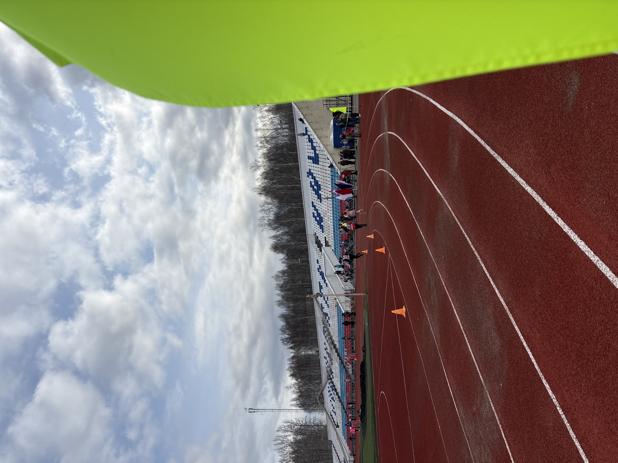 The image depicts an outdoor track and field stadium in Chaikovsky, Russia. The track is visible on the right side of the image, with white lane markings and orange cones positioned along the lanes. Seating is seen on the left, including spectator seating occupied by individuals, some of whom are holding flags. Behind the seating is a line of bare trees and a sky with cloud cover. A green object is partially obscuring the top left corner of the image.