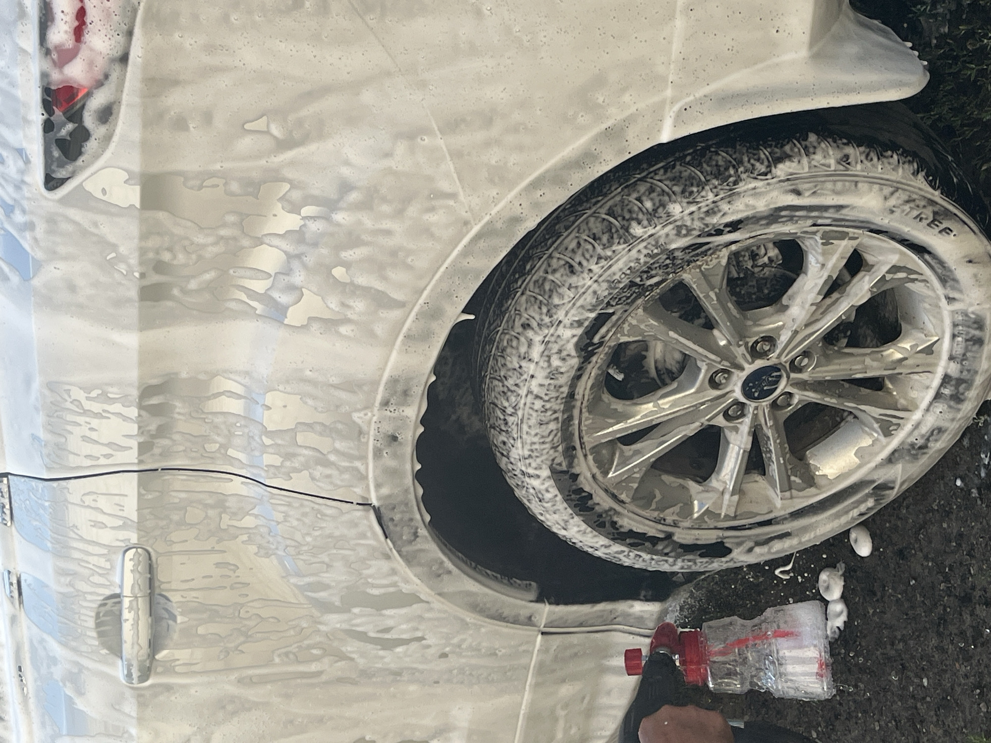 The image shows a close-up of a white car being washed. The car's exterior, including the wheel well and tire, is covered in thick white soap foam. A red and clear spray bottle, likely containing cleaning solution, is visible at the bottom right of the frame. A hand wearing a black glove is holding the spray bottle. The tire has some lettering visible, "STREE". The background appears to be dark, possibly asphalt, with some green foliage on the right side. The lighting suggests it might be daytime, but the weather is not clearly discernible due to the focus on the car and soap. The scene captures a moment of car washing activity.