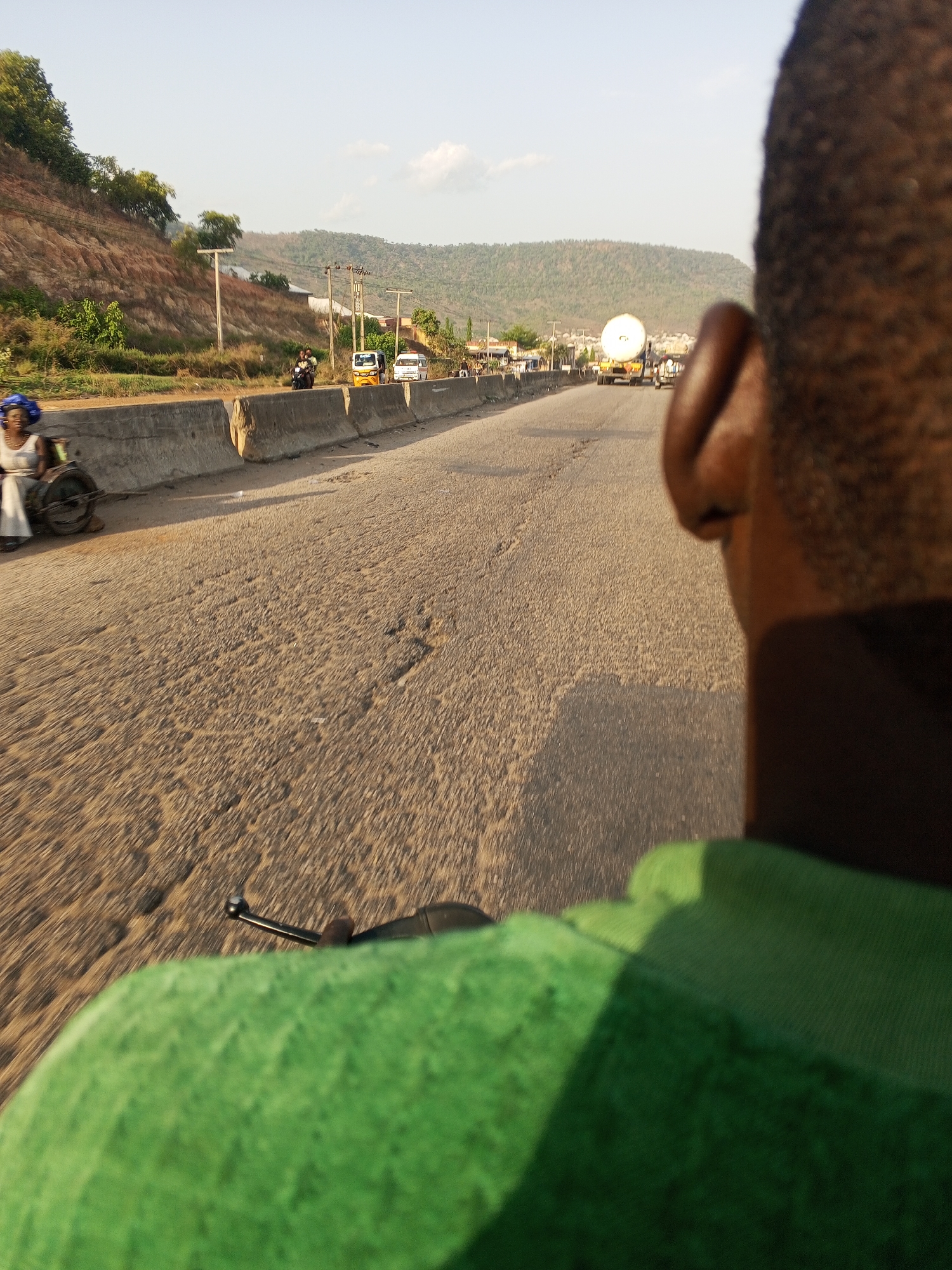 The image depicts a view from behind a person wearing a green shirt, likely on a moving motorcycle or vehicle, traveling along a road. In the foreground, the back of the individual's head and part of the green shirt are visible, along with a handlebar. The road appears cracked and worn. On the left side of the road, a concrete barrier separates the road from a shoulder where a woman in a white dress and blue headwrap sits in what appears to be a manual wheelchair. Further down the road, there are several vehicles including a tricycle taxi (keke), a white van, and a large truck. The background features a hilly landscape with vegetation and scattered buildings. The location is Lokoja, Nigeria, based on details consistent with roadside scenes in this city.