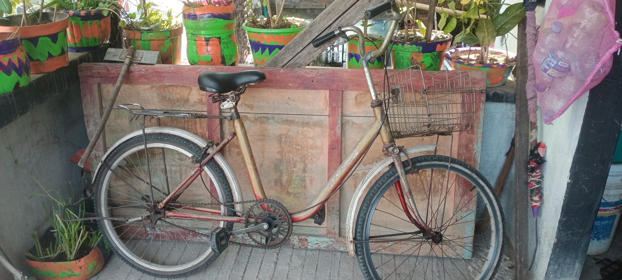 This is a medium-shot, eye-level, outdoor photograph of a vintage bicycle leaning against a wooden panel. The bicycle has a rusty metal frame, a black leather seat, a wire basket on the front, and fenders over both wheels. The front wheel is visible, as is part of the rear wheel and drivetrain. To the left of the bicycle, a garden tool with a metal head and wooden handle leans against the wooden panel. Behind the bicycle, several potted plants with colorful, hand-painted designs are visible on a ledge. The scene is likely set in a residential area in Semarang, Indonesia, given the style of the potted plants and the overall aesthetic. The ground beneath the bicycle appears to be paved with gray concrete tiles. A pink mesh bag filled with plastic bottles is hanging on the right side of the frame, near a white wall with black trim.