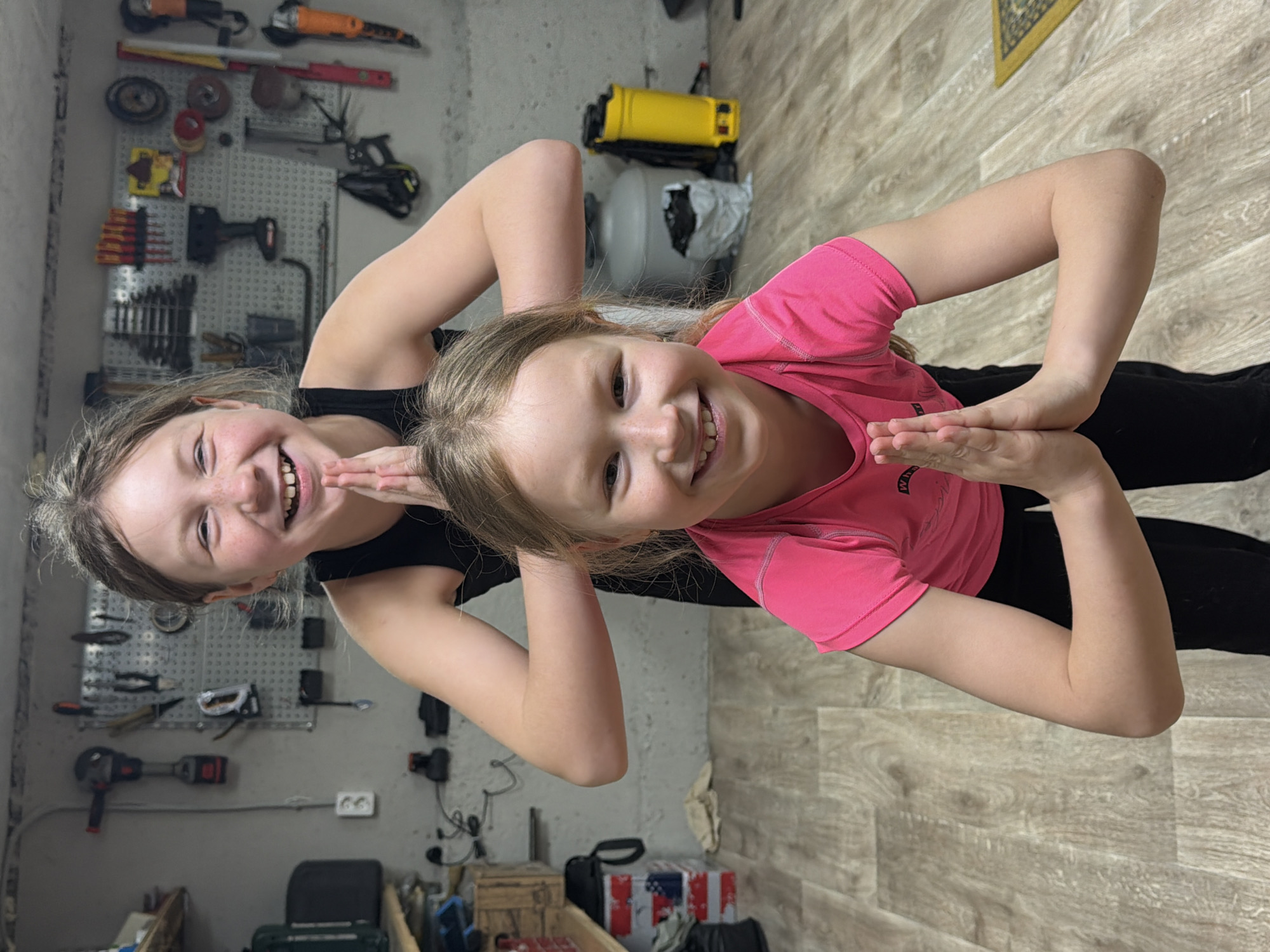 Two young girls are standing side-by-side, close together. One girl is wearing a pink shirt, and the other girl is wearing a black shirt. They are both smiling and have their hands pressed together in a gesture. The background appears to be a cluttered storage room or garage. There is a wall with tools hanging on it, various boxes and equipment on the floor, and flooring that mimics wood grain. The location is Dubovaya, Russia.