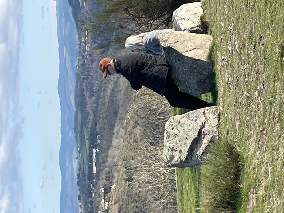 A daytime image depicts two individuals seated on large, weathered grey rocks atop a grassy hill overlooking a valley in Chambles, France. The male figure on the left is positioned with his back partially to the camera, wearing a brown baseball cap, dark sunglasses, a black quilted jacket, and dark trousers, with his arms crossed. To his right, a female with long blonde hair, dressed in a light grey top, is seated on an adjacent rock, also facing the distant landscape. The foreground consists of patchy green and brown grass with sparse low-lying vegetation. The background features a vast natural landscape under a partly cloudy blue sky, encompassing a valley with sparse deciduous trees, scattered residential structures, and several layers of distant blue-hued rolling hills extending to the horizon.
