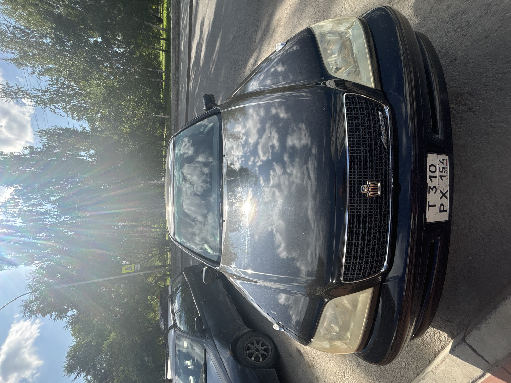 A black Toyota Crown sedan, model year unknown, is parked on a paved surface next to a curb. The car's front is facing the camera, and the hood reflects a cloudy sky. A license plate with the alphanumeric characters "T310 PX 154 RUS" is affixed to the front of the vehicle. In the background, several deciduous trees line a road, and power lines are visible in the distance against a bright, sunlit sky. The overall lighting suggests it is daytime. The image appears to have been taken on a street in Novosibirsk, Russia.