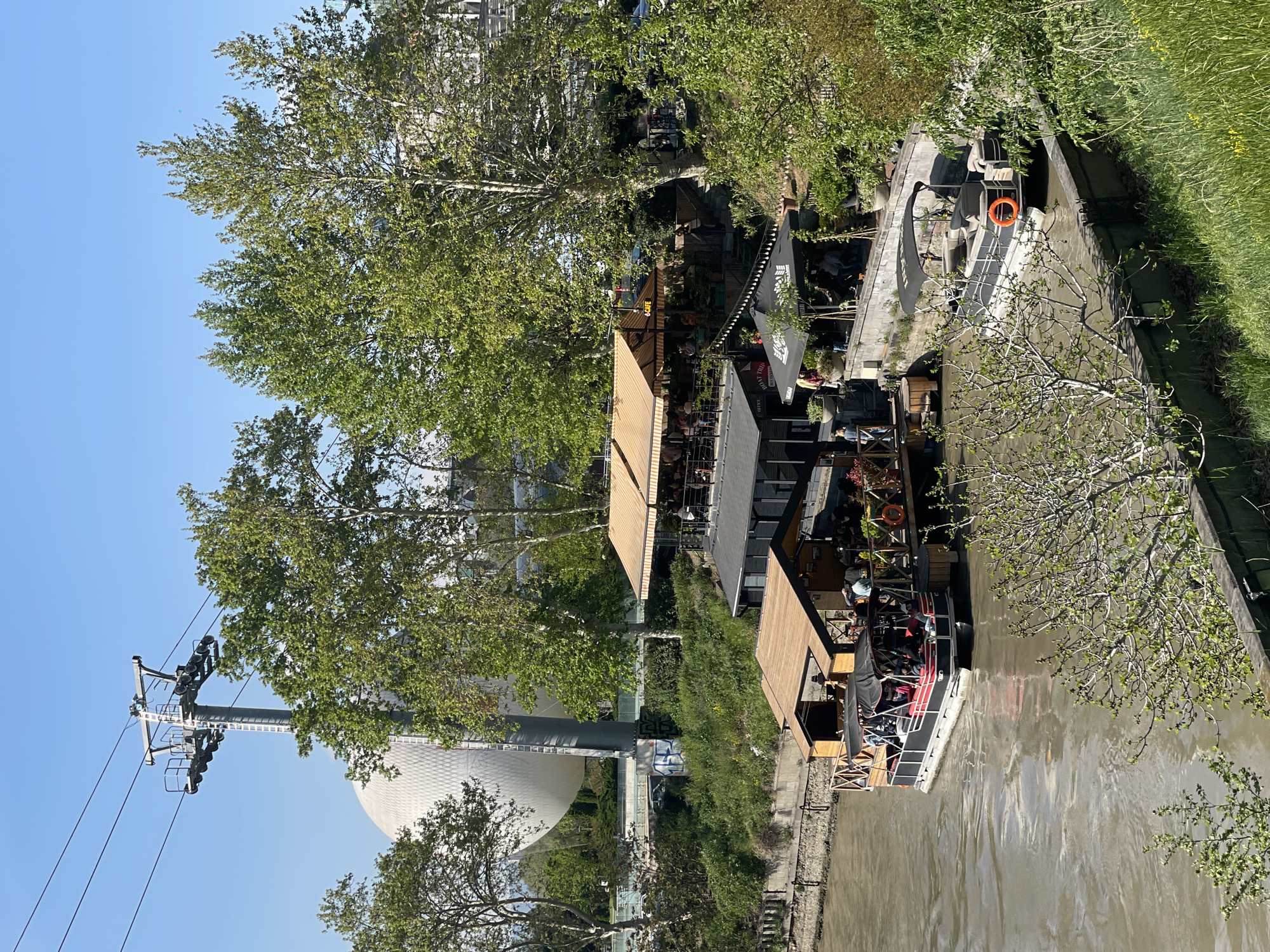 The image captures a vibrant riverside scene in Tbilisi, Georgia, on a clear, sunny day, likely during late morning or afternoon. The setting features a river flowing through an urban area, flanked by green banks and numerous trees.
Along the left bank, several open-air cafes or restaurants are built, featuring wooden structures and outdoor seating areas. Many patrons are visible, relaxing at tables under awnings and large umbrellas. One black umbrella prominently displays white text that appears to read "THE CHAIR," and another wooden sign above an entrance shows "COFFE" or "CAFE." These establishments create a lively atmosphere by the water.
The river itself is wide with calm, murky brown water. Several boats are present; one larger, flat-topped boat or floating platform, possibly an extension of a restaurant, is moored near the bank and features an orange lifebuoy. Further along the river, a narrower boat with red seating is visible, implying recreational activities or river transport. The opposite bank of the river is grassy and dotted with trees and shrubs.
In the background, on the left side of the frame, iconic Tbilisi landmarks are visible. A large, white, textured dome-shaped building stands out, likely the Presidential Palace or a similar structure near Rike Park. Adjacent to it, a tall metal pylon with multiple cables extends towards the sky, indicating the Narikala Cable Car system. Lush green foliage from various trees frames the scene, providing shade and natural beauty to the urban landscape. The overall impression is one of leisure and activity in a scenic city location.