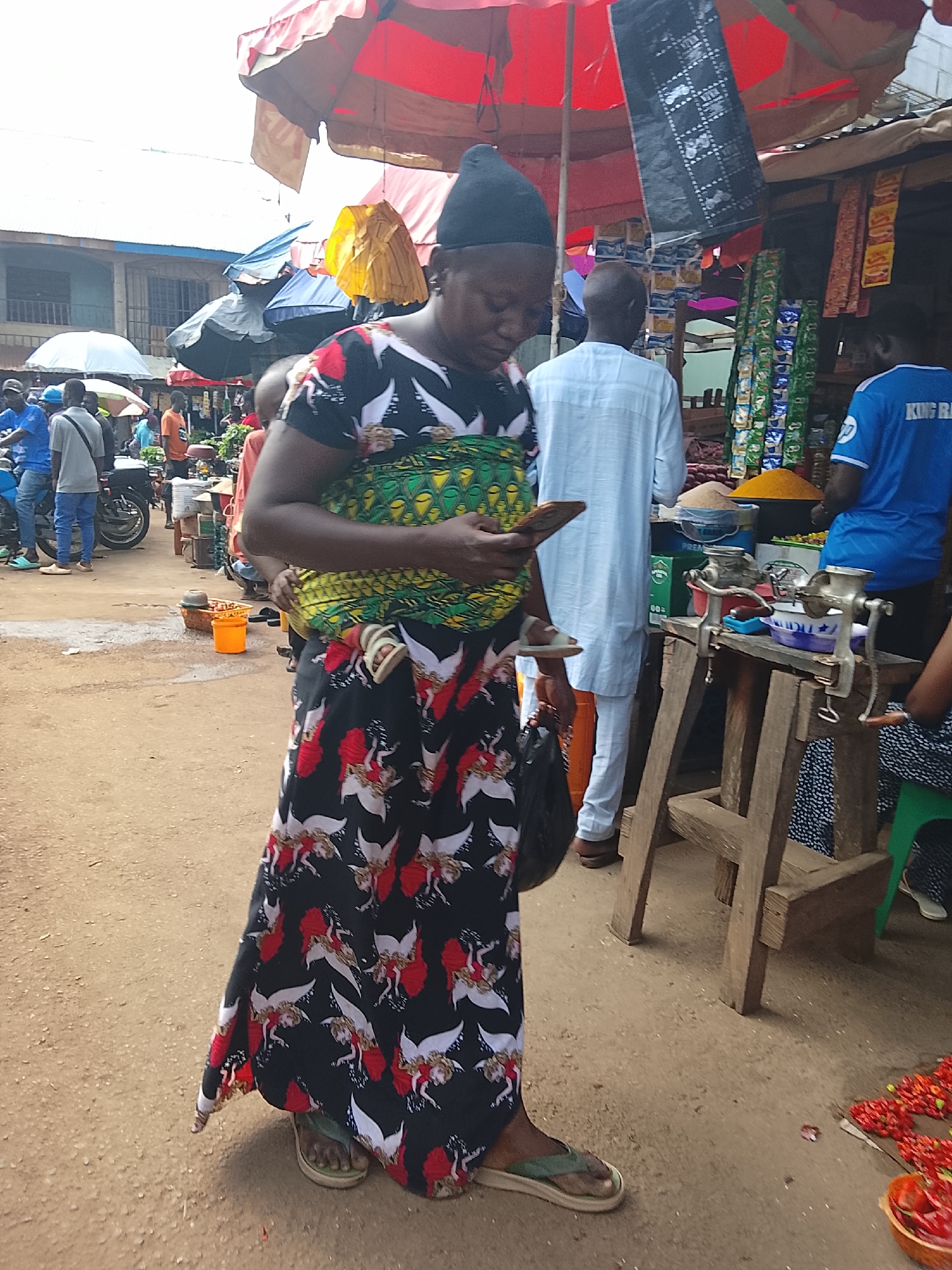 A medium shot captures a busy outdoor market scene in Akwanga, Nigeria. The focal point is a woman in the foreground, wearing a black headscarf, a black and white patterned top with red accents, and a long, flowing black skirt adorned with a repeating pattern of red figures and white winged creatures. She is looking down at a smartphone in her right hand. To her left, a man in a white shirt and pants stands facing away from the camera. Behind them, stalls are set up with various goods, including sacks of grain and colorful items on shelves. In the background, several people are visible, some standing near motorcycles and others browsing at the market stalls. Red and yellow umbrellas provide shade. The ground is a dusty, sandy surface. To the right, a wooden table holds two manual meat grinders, and a bowl of red peppers is on the ground. Another person in a blue shirt is visible behind the table.