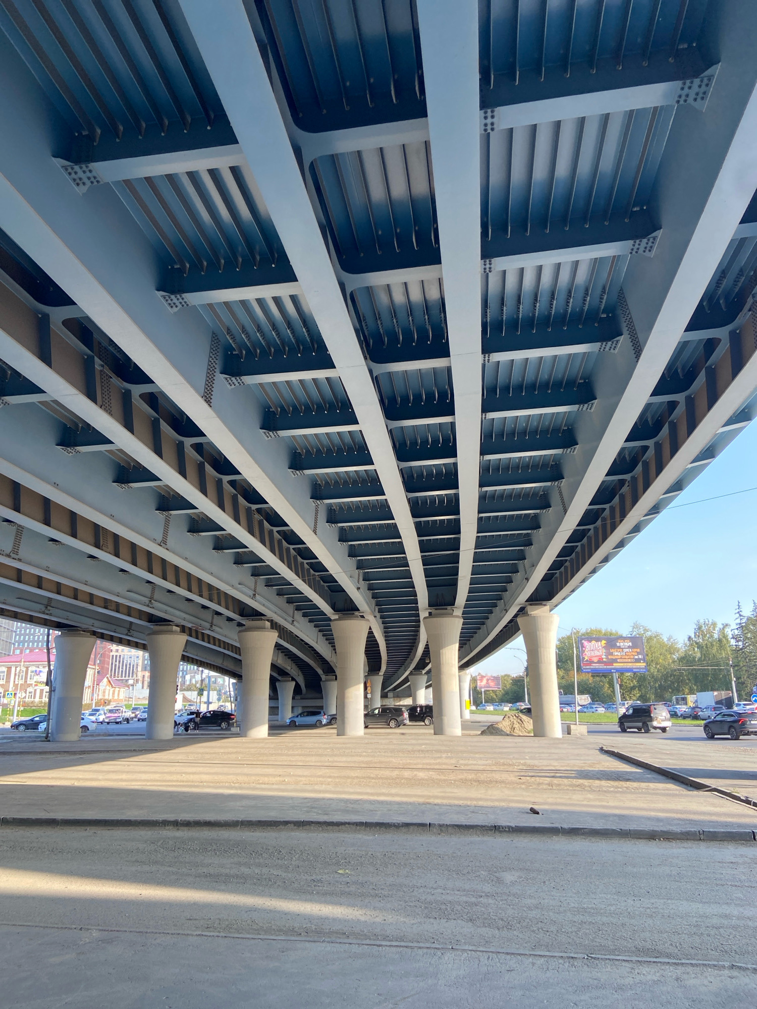 This is an outdoor, low-angle, wide shot of the underside of a large, modern overpass on a clear, sunny day. The overpass is made of light gray steel beams and girders, with a light blue corrugated metal ceiling. The structure is supported by numerous thick, white concrete pillars. The ground beneath the overpass is paved with concrete and shows some shadows. In the background, several cars are driving on a road. On the right side of the background, there is a billboard with Russian text and images. Trees and some buildings are also visible in the distance. The scene appears to be in an urban or semi-urban area. Based on the billboard, which advertises a performance on October 16th, and the architectural style of the overpass, this location is likely in Novosibirsk, Russia. There are no people clearly visible in the frame.