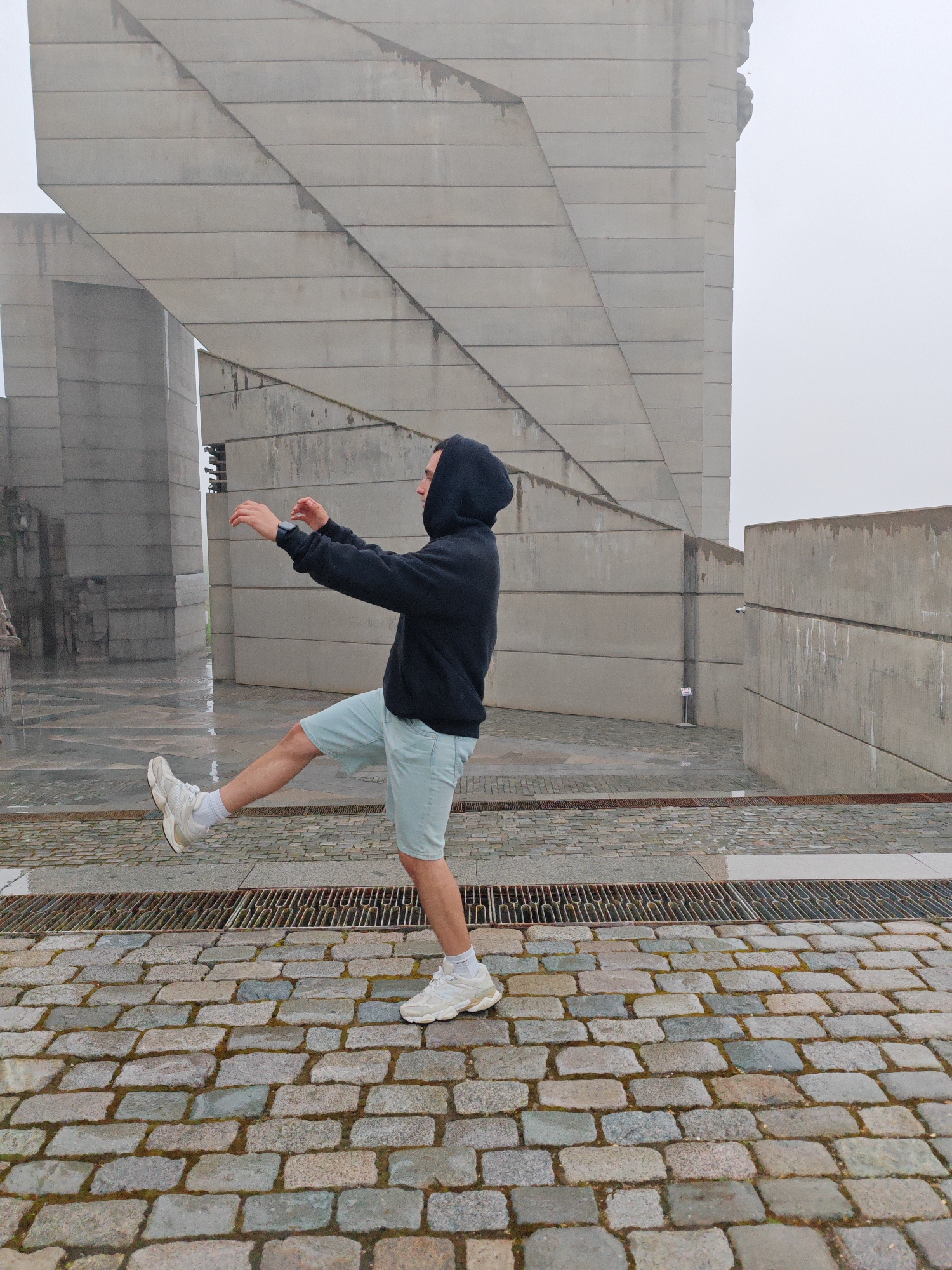 A young man stands on a cobblestone surface in front of a large, modern, concrete monument in Shumen, Bulgaria. The monument consists of large, blocky, angled structures, typical of socialist-era architecture. The sky is overcast with a thick fog obscuring details in the background. The man is wearing a dark hooded sweatshirt and light-colored shorts. He is holding his arms out and is in the process of lifting one leg. He is wearing white sneakers and socks. A metal grate runs between the person and the monument.