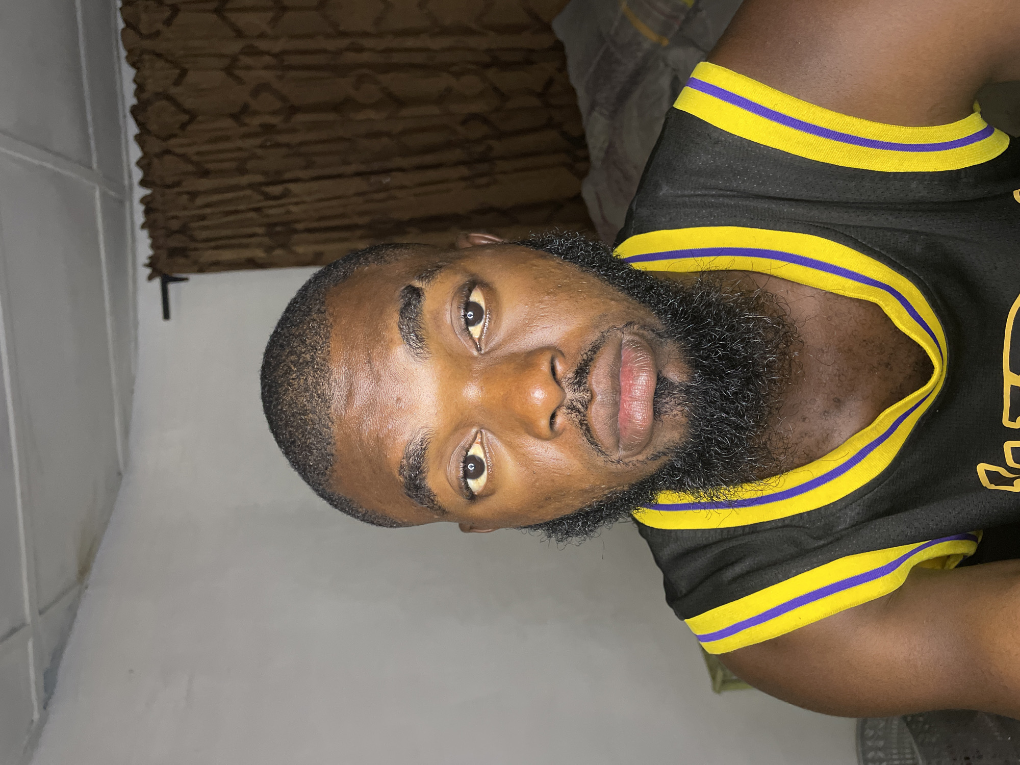 A close-up, frontal portrait of a Black man with a short haircut and a full beard is shown. He is wearing a black sleeveless jersey with yellow and purple trim around the neckline and armholes. The background consists of a light-colored wall with a brown curtain to the right, and a ceiling with visible panels and a stain above. The man's expression is neutral, and his eyes are looking directly at the camera. The lighting suggests an indoor setting, possibly at night or in a dimly lit room. The image does not provide any specific geographical indicators that would place it in Jos, Nigeria.