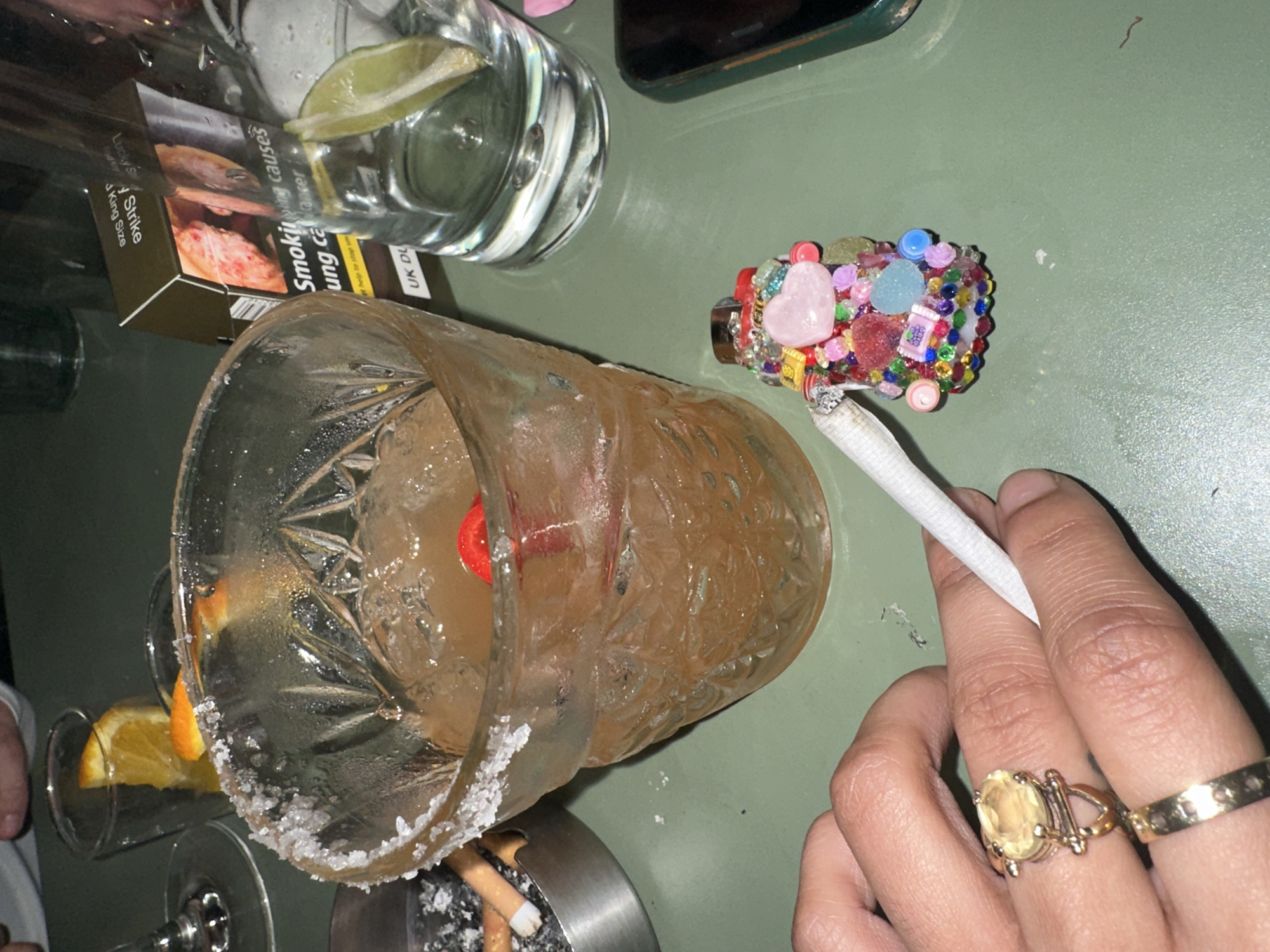 The image depicts a scene on a green table, potentially in a London establishment. On the table is a cocktail in a glass with a salted rim, a cigarette holder adorned with colorful decorations holding a cigarette, a pack of "Lucky Strike" cigarettes (UK DU version), a glass of water with a lime wedge, a mobile phone, an ashtray with cigarette butts, and a hand holding a cigarette. The hand features two gold rings. A second cocktail glass can be seen with an orange garnish.