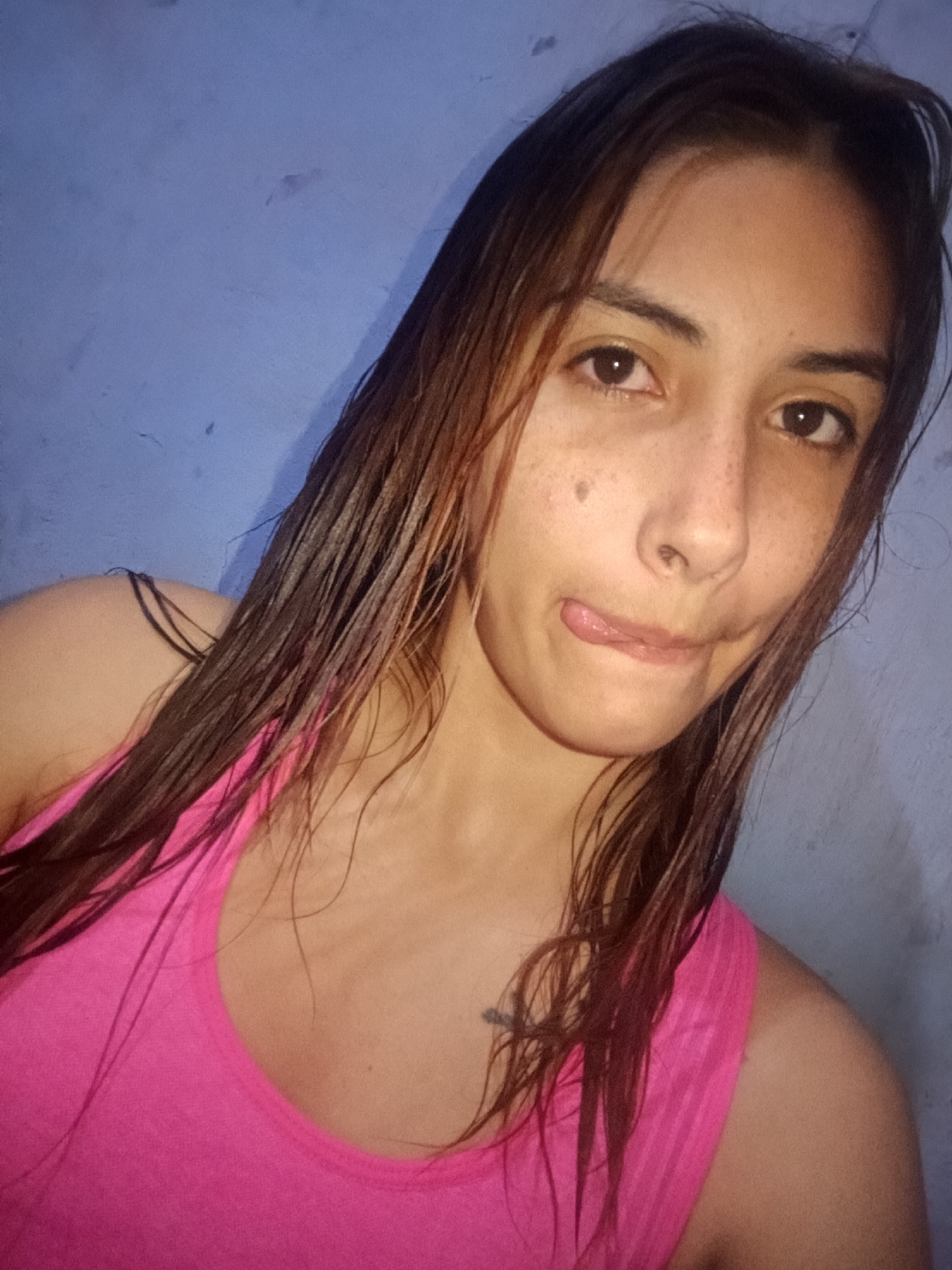 A young woman is captured in a close-up, informal portrait, likely a selfie. She has long, dark, and visibly wet hair that cascades over her shoulders and chest. She is wearing a bright pink tank top with thin straps, one of which is partially visible over her left shoulder.
Her face is turned slightly to the right, and she has a playful expression, with her tongue peeking out and her lower lip gently bitten. Her brown eyes are open, and her face shows natural features, including several freckles across her nose and cheeks, and a small mole on her right cheek below her eye. The background is a plain, textured light blue wall, suggesting an indoor setting. The lighting appears soft and somewhat dim, casting subtle shadows. The overall impression is casual and lighthearted. No text is visible in the image. The location is Juan Griego, Venezuela.