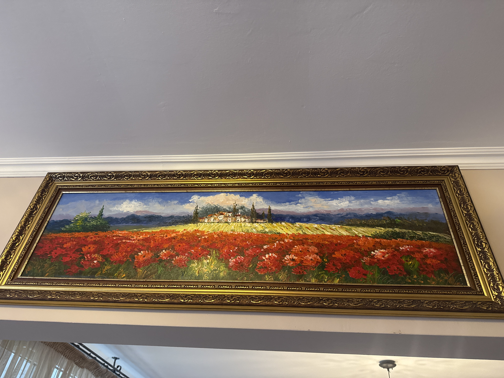 A landscape painting in an ornate gold frame hangs on a light-colored wall. The painting depicts a field of red flowers in the foreground, leading to a patch of yellow crops and a village or small town in the middle ground. A blue sky with white clouds is visible above. Parts of a room interior are visible in the lower part of the frame, including a light fixture and a curtain. The image does not contain information about Plovdiv, Bulgaria.