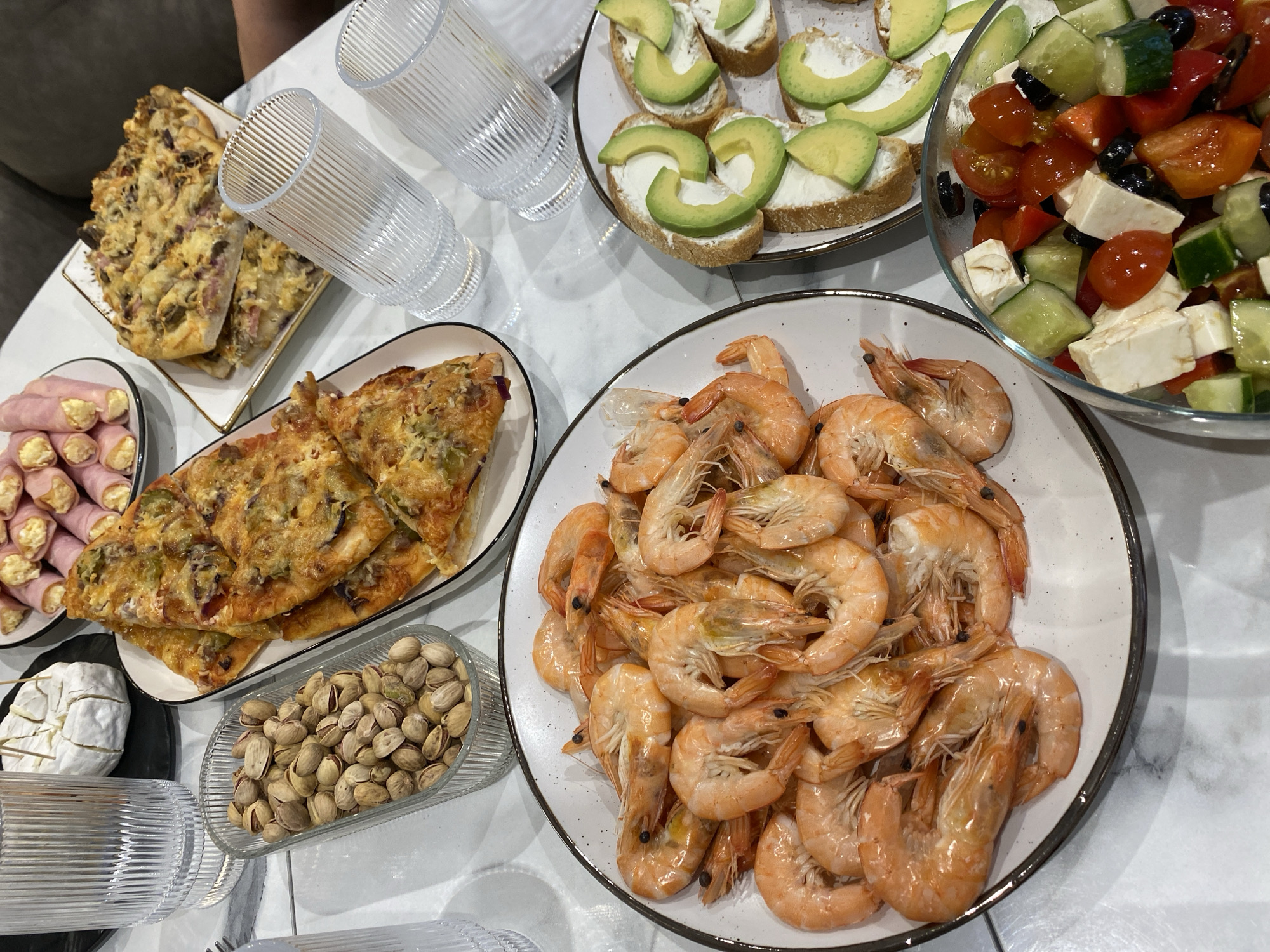 A well-lit, high-angle shot showcases a spread of food on a white marble-patterned table. The scene includes a variety of appetizers and dishes, suggesting a social gathering or meal setting.
Prominent in the foreground is a large white plate filled with cooked shrimp. Adjacent to the shrimp is a clear dish containing pistachios. To the left of the pistachios is a black plate with a wheel of brie and a plate with sliced ham rolls. Another plate holds slices of pizza or flatbread.
At the top of the image, there is a plate with bread slices topped with cream cheese and avocado slices. A glass bowl filled with a salad comprised of tomatoes, cucumbers, feta cheese, and black olives completes the assortment of food. Three clear, textured glasses are placed near the center of the table.
The scene is indoors. The city of Izhevsk, Russia, is not discernible from the given visual information.