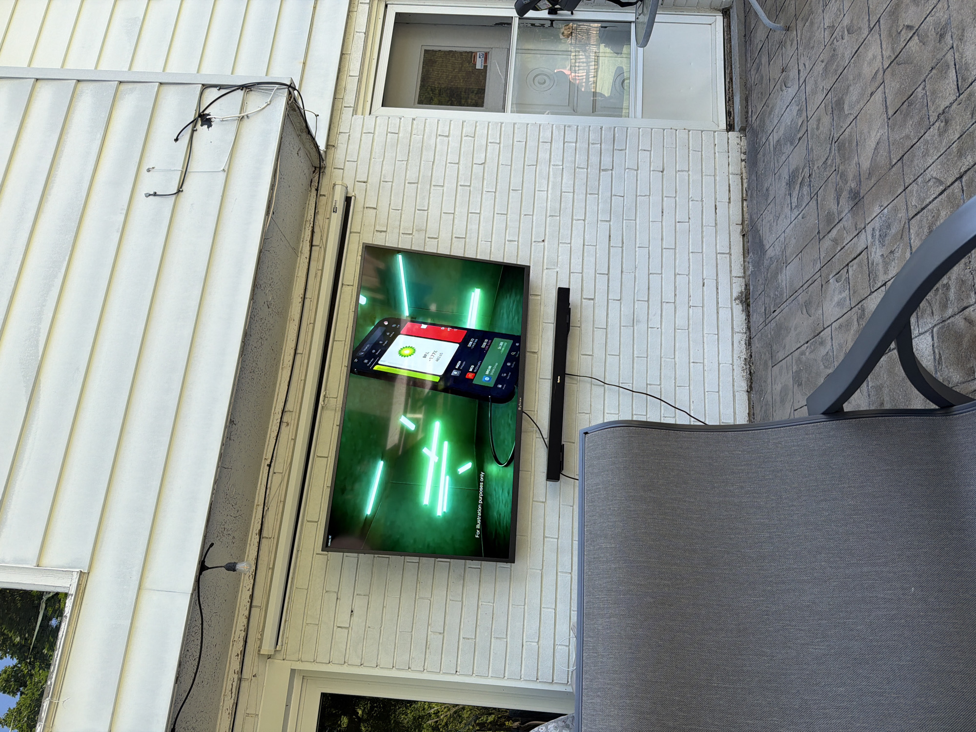 An outdoor television and soundbar are mounted on a white brick wall, part of a residential building's exterior, likely a patio or deck area. The TV displays a vibrant digital image featuring glowing green rectangular shapes and a stylized smartphone interface, with the text "For illustration purposes only" visible at the bottom. Smaller text and numbers like "89.1", "-1.77%", and "483.05" are visible on the simulated phone screen. A brand name "SYINXI" is faintly visible on the TV's right frame.
To the left of the white brick wall, an adjacent section of the building features white horizontal siding, with various black and white electrical wires and a single light fixture attached. Above the TV and brick wall is a white-framed glass door or large window. The ground area is paved with dark grey, textured paver stones. In the foreground, a portion of a grey outdoor chair, including its armrest and back, is visible. The scene is brightly lit, suggesting daytime. No people or specific activities are occurring. The setting indicates an outdoor entertainment space.