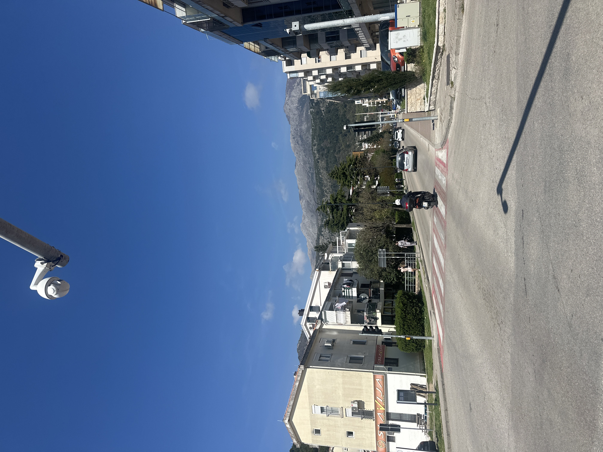 The image captures a street scene in Budva, Montenegro, on a sunny day. The sky is clear blue. The shot is taken from a slightly elevated position, possibly near a building or an elevated part of the street. Buildings are visible on both sides of the street, showing a mix of residential and commercial structures. In the distance, there are mountains. A street with a crosswalk is in the midground. There is a motorcyclist on the crosswalk. There are cars in the distance on the road. A security camera on a pole is visible in the lower-left corner.