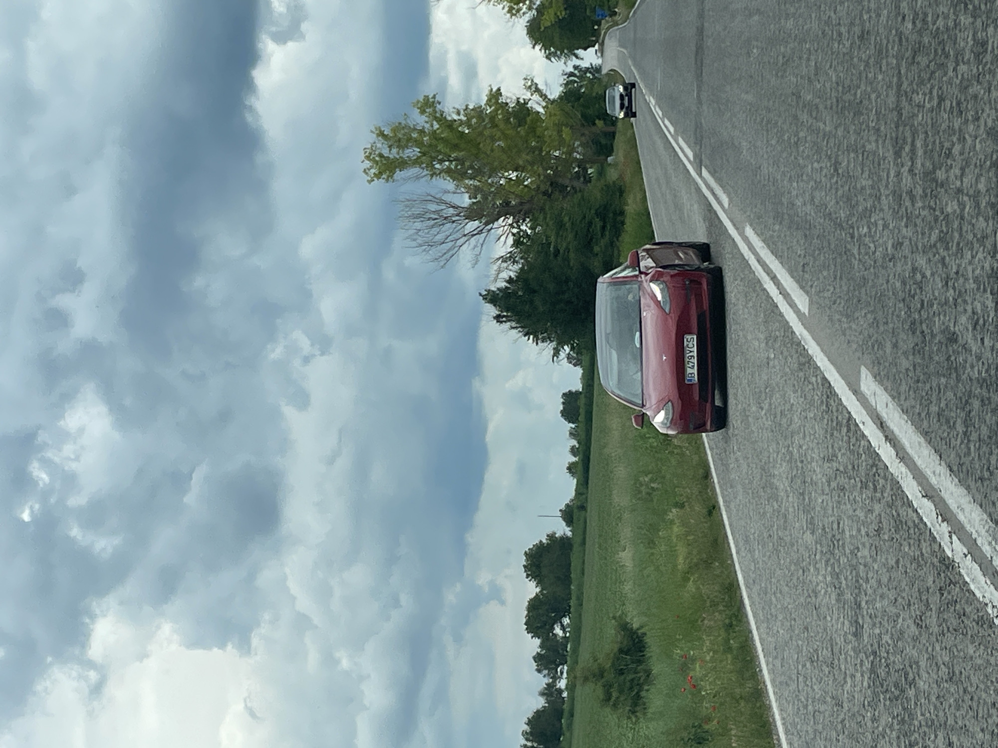 A landscape photo depicts a two-lane road with a red car parked on the right shoulder and a silver car in the distance. A lush green field borders the roadside, transitioning into a dense tree line. The sky above is filled with textured clouds. The registration plate on the car nearest to the viewer is "B 479 VCS". The location is potentially in or near Kamenar, Bulgaria.