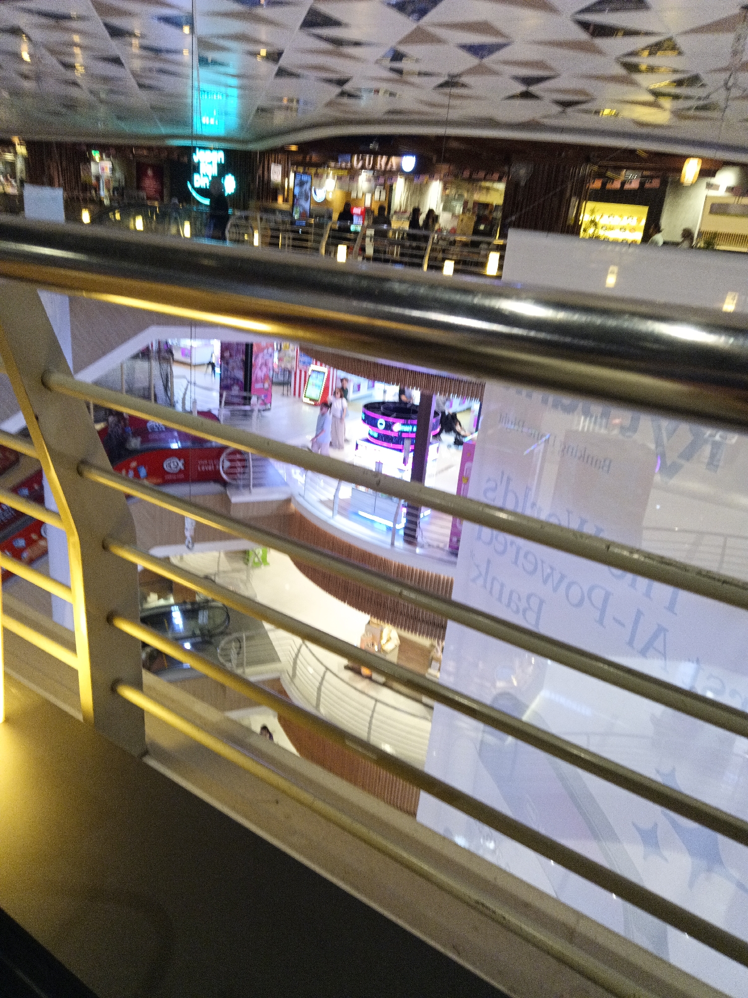 This image shows an interior view of a multi-level shopping mall, likely in Kuala Lumpur, Malaysia, looking down from an upper floor. The foreground features a metal railing and a balcony or walkway. Below, the mall's interior is visible, with various shops, escalators, and people. In the middle ground, there is a red advertisement featuring the logo "EX". Further down, a large banner or advertisement is displayed, with text that appears to read "The Power-IA Bank" in a mirrored fashion. The ceiling features a geometric patterned design.