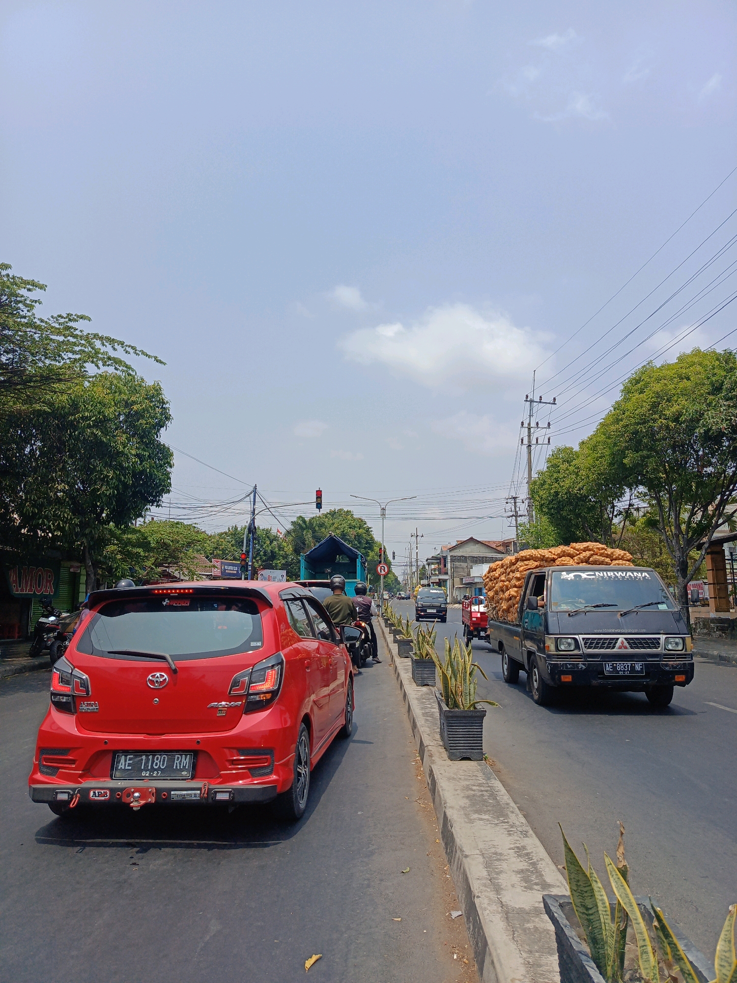 This is a vertical, eye-level, medium shot of a street scene in Sukowinangun, Indonesia. The street is busy with traffic, including a red car in the foreground, a black truck loaded with sacks of produce, and several motorcycles. The sky is partly cloudy, and trees line the street. A traffic light is visible in the distance. The road is paved, and there is a concrete median with potted plants. Buildings and businesses are visible on either side of the street. The red car has a license plate that reads "AE 1180 RM." The truck has a license plate that reads "AE 8837 NF." A person wearing a helmet is riding a motorcycle behind the red car.