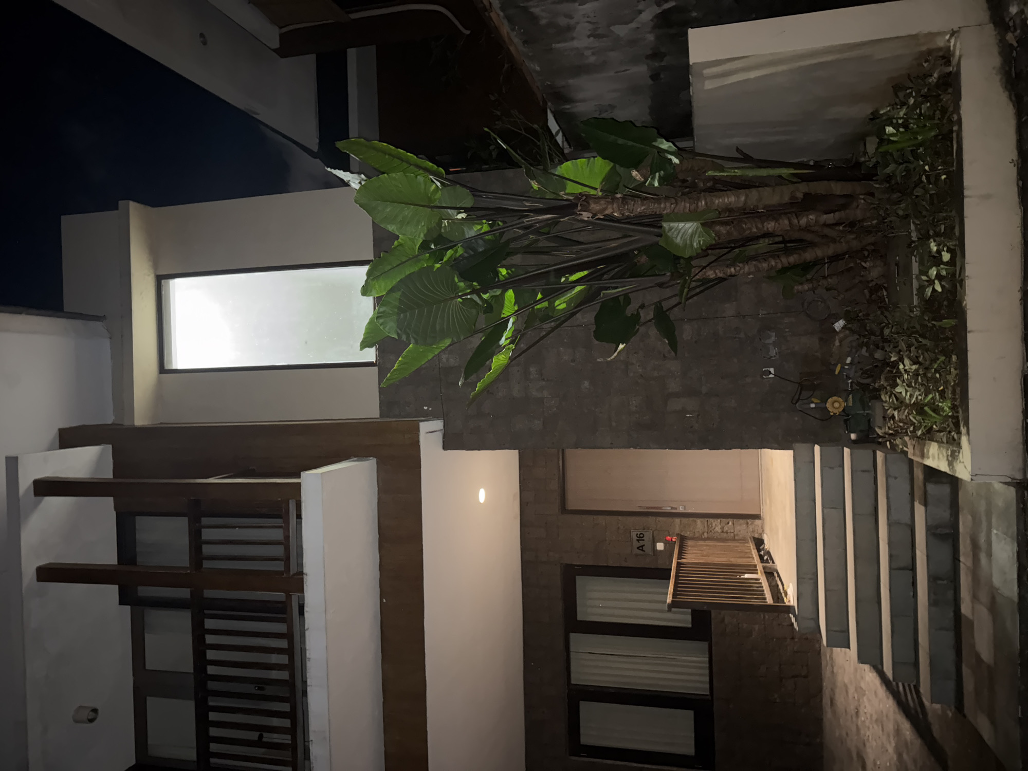 The scene depicts a multi-story building exterior in Kuta Selatan, Indonesia, under nighttime conditions. The building features a combination of modern and rustic elements, including stone walls, white pillars, wooden accents, and a large window on the upper level, which emits a bright light. Lush, tropical foliage, potentially decorative, is situated on the roof level of one section of the building. A set of concrete stairs leads up to an entranceway, and a partial view of an exterior window is visible, indicating possible residential or commercial units. "A 16" is displayed next to a red decoration on the outside.