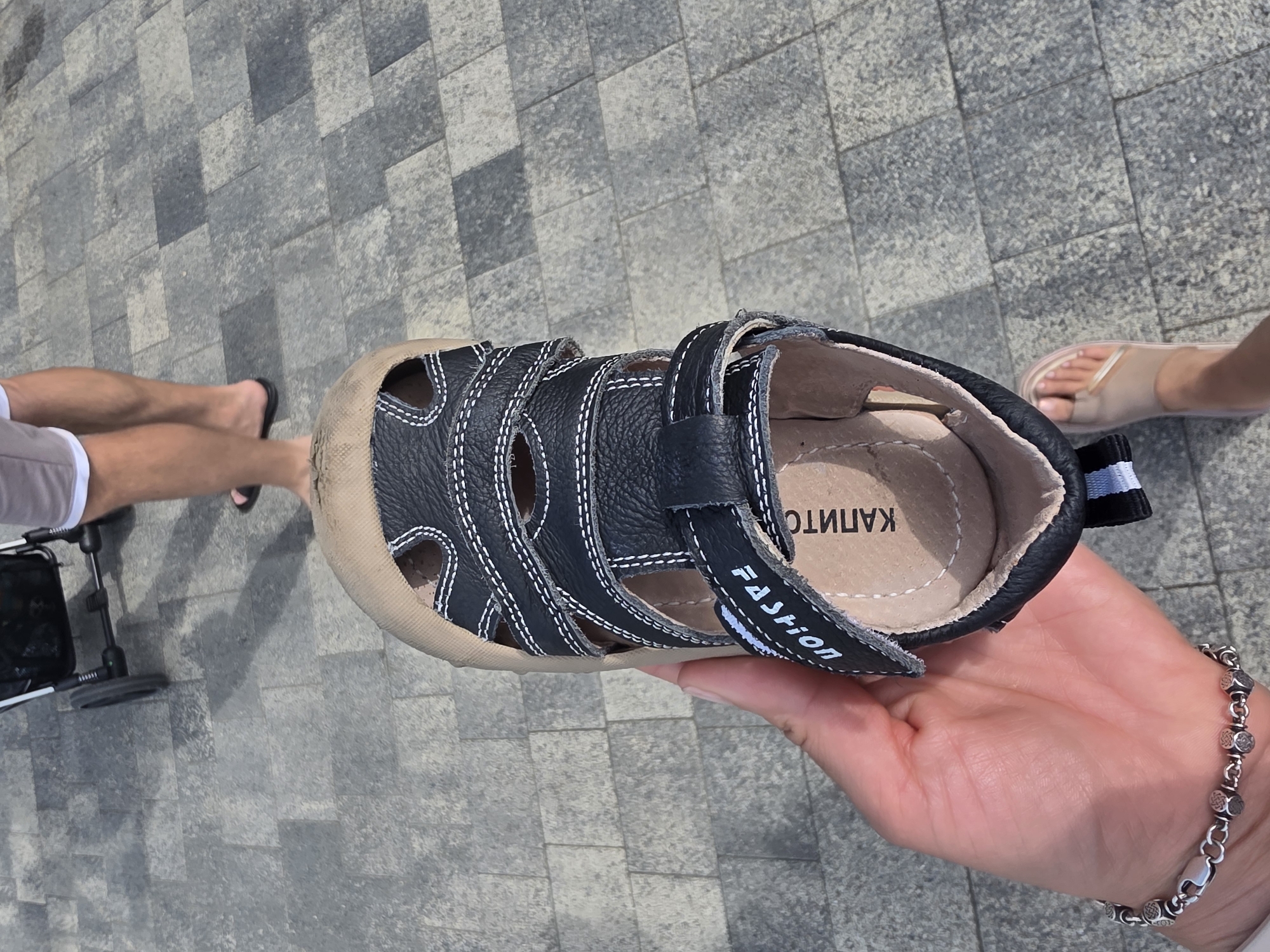 A person's hand holds a black sandal with white stitching and a tan interior, over a grey paved surface. The sandal has two velcro straps across the top and a strap with "FASHION" printed in white. The words "КАПИТО" are visible on the insole. In the background, two pairs of legs are visible walking on the same paved surface. One pair of legs wears grey shorts and flip-flops, and the other pair of legs is clad in a skirt and sandals. A baby stroller is also partially visible in the background. The setting is an outdoor paved area, consistent with a public walkway or street in Sochi, Russia.