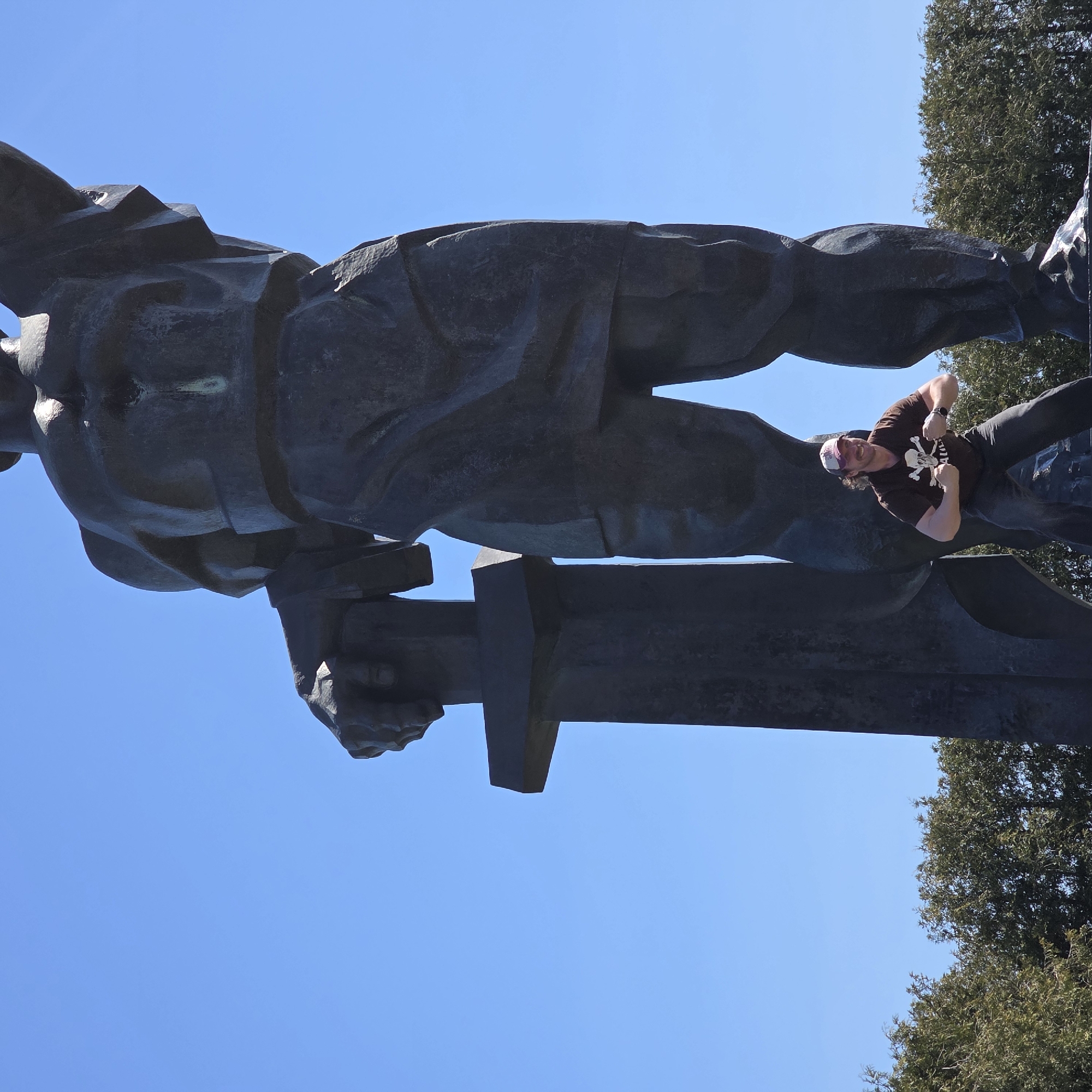 A man is hanging upside down on a statue in Свіслацкі сельскі Савет, Belarus. The statue is dark, possibly bronze, and depicts a figure holding a sword. The man is wearing a dark t-shirt with a skull and crossbones design, dark pants, and a cap. He has his arms bent, fists near his chest, and is smiling. The sky is blue in the background, with trees visible in the upper right corner.