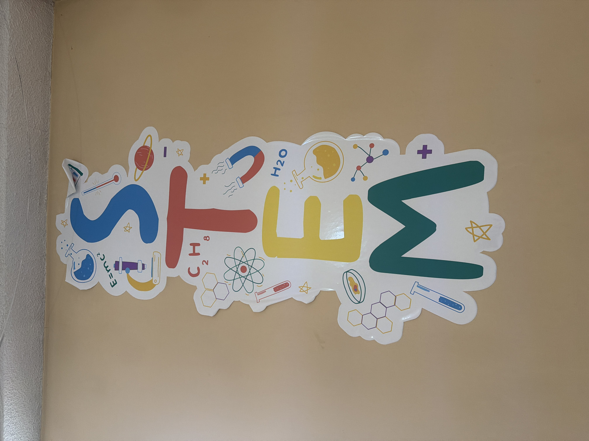 A vibrant "STEM" sign is prominently displayed on a plain, light beige wall. The acronym is rendered in large, colorful letters, with "S" in blue, "T" in red, "E" in yellow, and "M" in green, all outlined with a white border.
Each letter is adorned with various scientific and mathematical illustrations. The blue "S" features a beaker, a thermometer, a ruler, a stylized planet with rings, and stars. Around the red "T" are a horseshoe magnet, the chemical formula "H2O" (water), a beaker with bubbling liquid, a molecular structure, "C2H8" (likely an organic compound), and an atomic model. The yellow "E" showcases hexagonal chemical structures, another atomic model, and a test tube. The green "M" includes more hexagonal chemical structures, a test tube with blue liquid, and stars. Other elements scattered across the sign are plus signs, stars, and the famous equation "E=mc^2".
The scene is an indoor setting, likely an educational environment such as a classroom, laboratory, or school hallway in Sofia, Bulgaria, given the thematic content. No people are visible, and no activity is taking place; the image captures a static, decorative, and educational display. A portion of a white vertical structure, possibly a wall corner or door frame, is visible on the far left. The colorful and illustrative design aims to be engaging and inspiring for subjects related to Science, Technology, Engineering, and Mathematics.