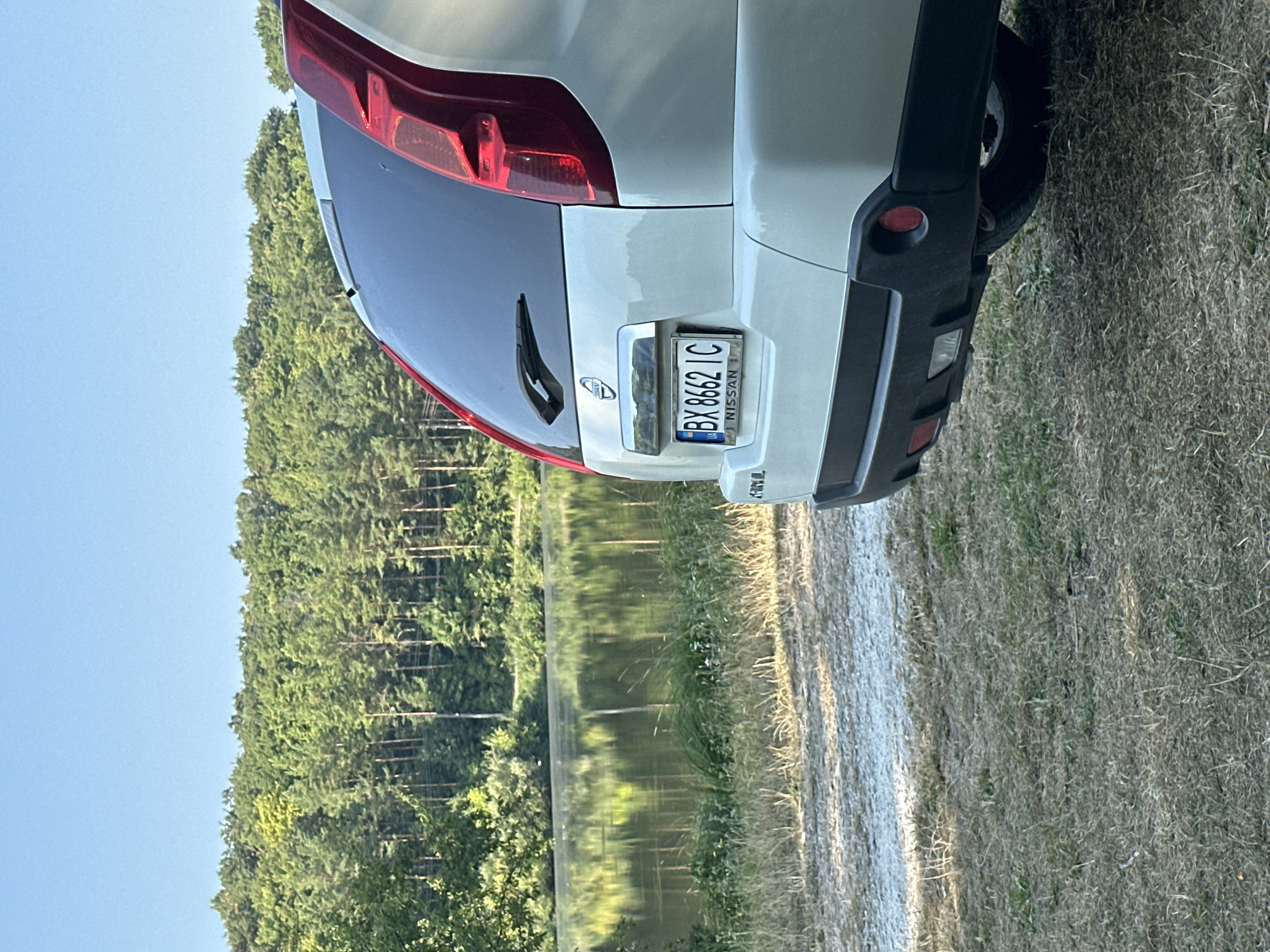 The image shows the rear of a white Nissan SUV parked on a dirt and grass roadside. The car's license plate reads "BX 8662 IC". In the background, a calm lake reflects a dense forest of green trees under a clear blue sky. The scene appears to be a natural, outdoor setting. The provided location of Makiv, Ukraine, suggests this is a rural or semi-rural area.
