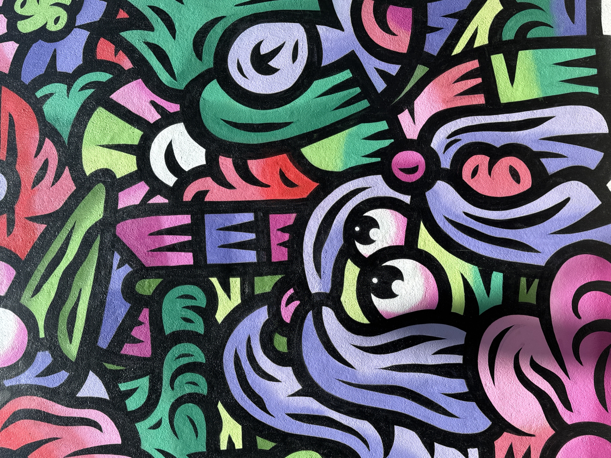 This is a detailed close-up of a vibrant, abstract street art mural or graffiti located in Sofia, Bulgaria. The artwork features a dense, interlocking pattern of organic and geometric shapes outlined in thick black lines. The dominant colors include various shades of green, purple, pink, red, and light yellow-green, with some areas exhibiting subtle color gradients. Towards the bottom center, two large, stylized, cartoon-like eyes with prominent white irises and black pupils are visible, belonging to a creature, possibly a dog or mythical being, characterized by flowing purple 'fur' or 'hair' and a small pink snout-like feature. Surrounding this central figure are numerous abstract forms suggesting foliage, stylized flowers, and geometric patterns. The mural is painted on a textured wall surface, likely concrete or stucco, and is illuminated by bright, even lighting, creating minor shadows that add depth to the painted forms.