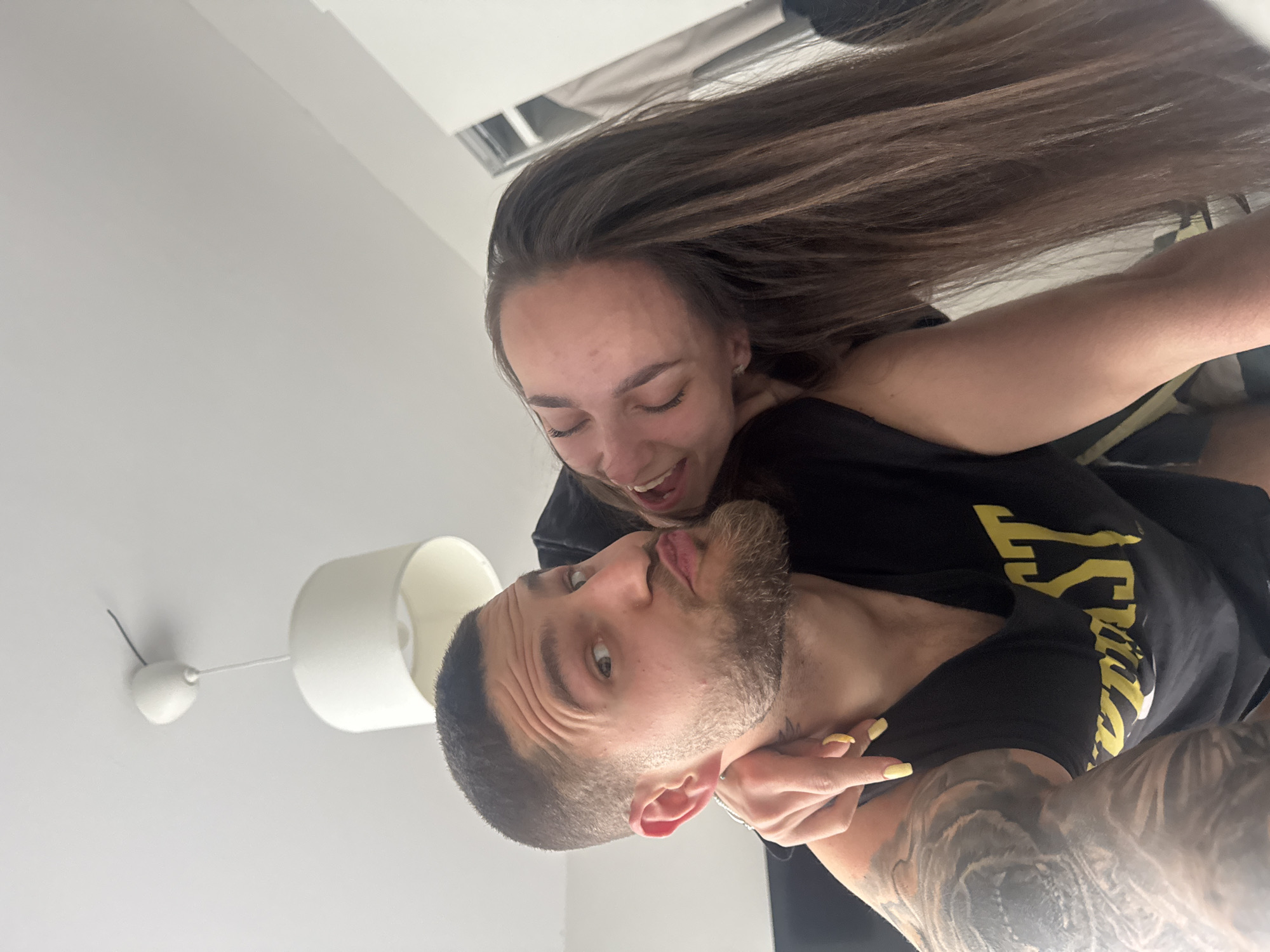 A man and a woman are in a playful interaction. The man has a short haircut, beard, and a tattoo on his arm. He is wearing a black t-shirt. The woman has long brown hair and is smiling with her eyes closed. She is also wearing a black top and has her arm around the man's neck. The background is a light-colored wall with a white ceiling fan. A white lampshade is also visible in the background. The location appears to be indoors, possibly in a room in an apartment. There is not enough information to identify the specific location in Sofia, Bulgaria.