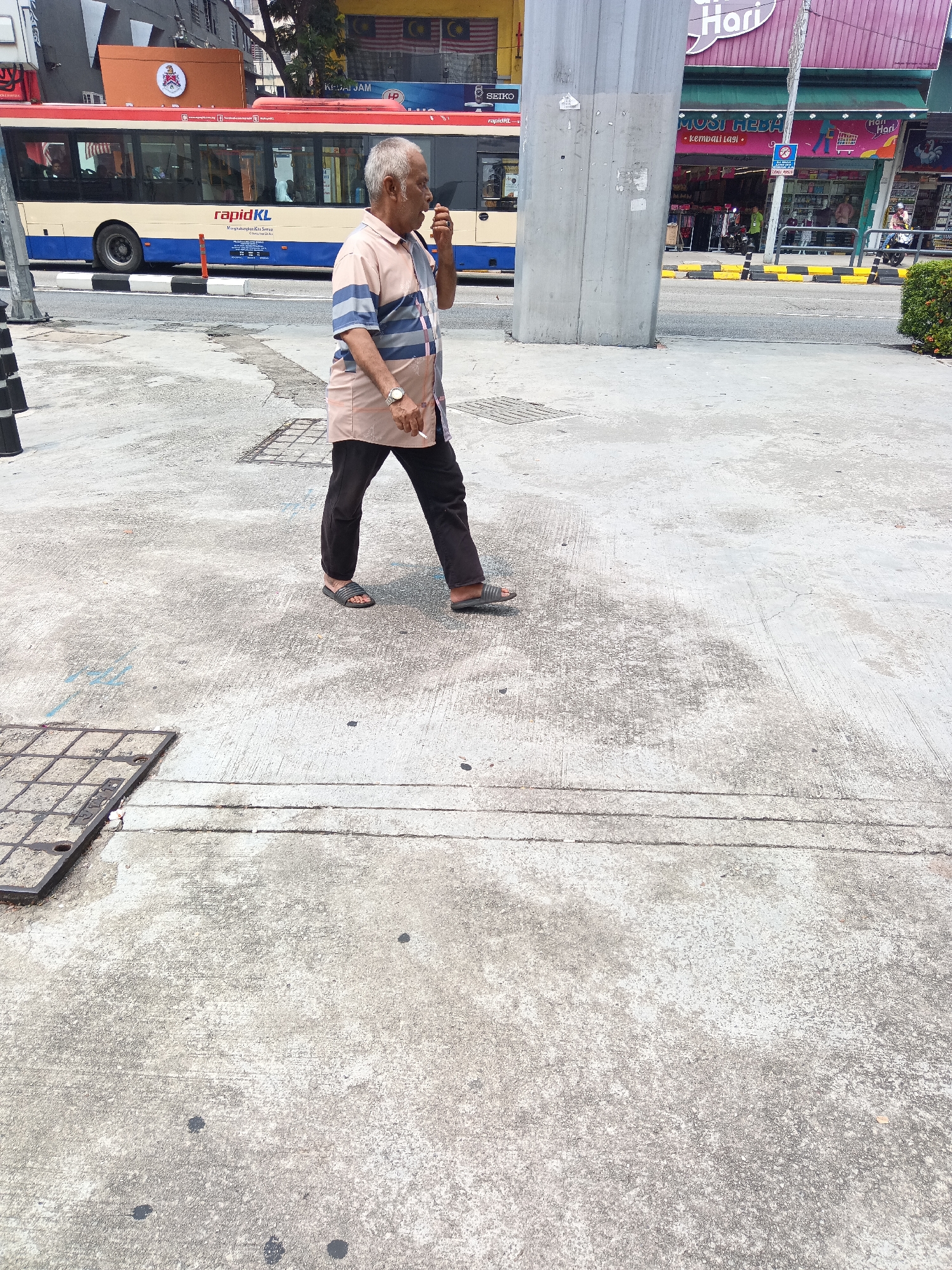 An elderly man with graying hair walks along a concrete sidewalk in Kuala Lumpur, Malaysia, with a red and cream-colored bus labeled "rapidKL" passing behind him. The man is wearing a short-sleeved, striped button-down shirt and dark pants, and appears to be smoking. He is wearing dark sandals. The sidewalk is cracked and weathered, and there is a metal grate visible on the left side of the frame. In the background, a building with a green awning and various signs can be seen, including one that reads "HARI HARI" and another that says "SEIKO". The scene suggests an urban environment with public transportation and commercial activity.