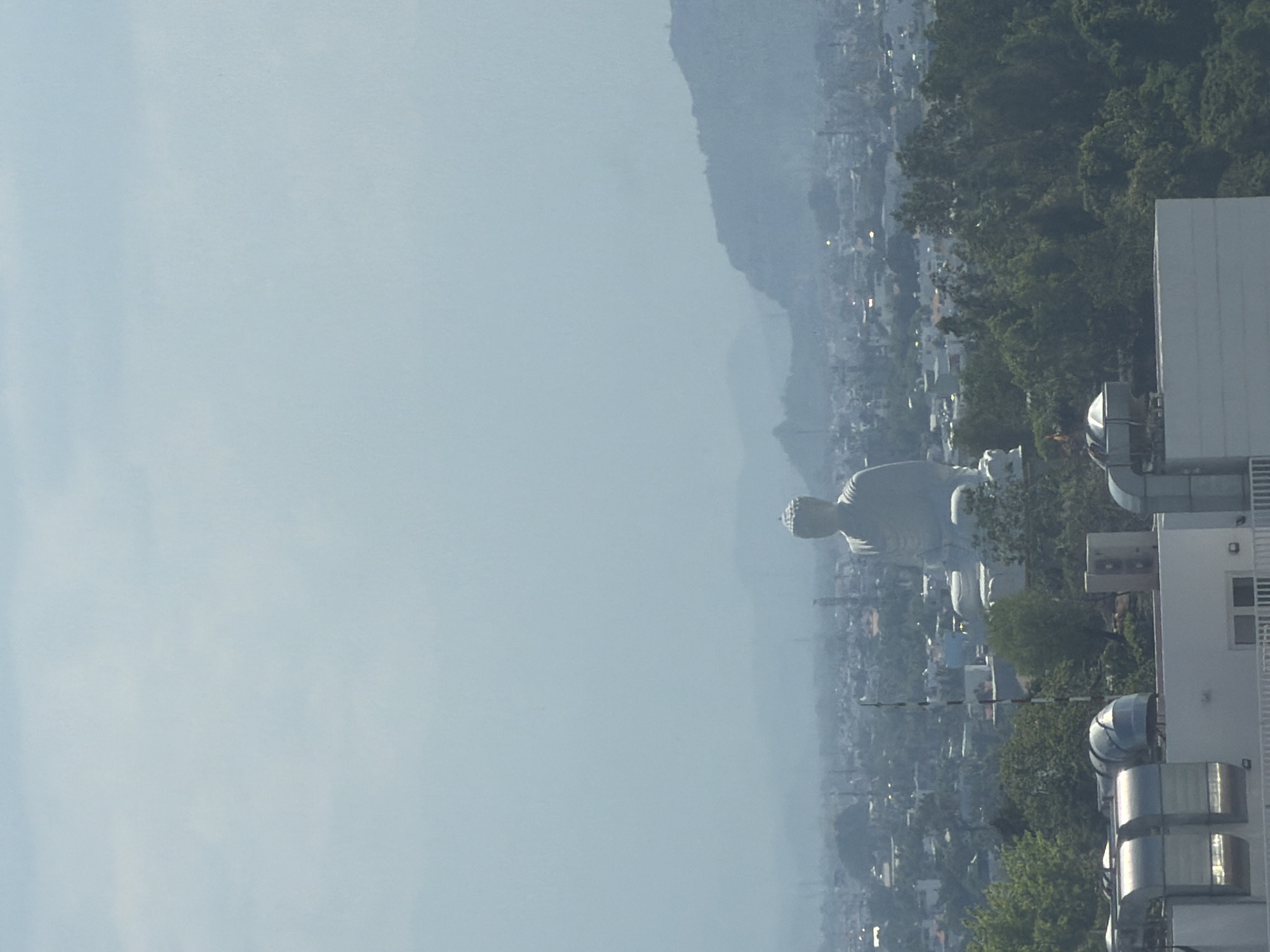 The image captures a panoramic view of a city with a colossal statue of a seated Buddha as the prominent focal point. The statue, rendered in a light, monolithic material, is situated on an elevated platform, commanding the cityscape. The Buddha is depicted with a serene expression, hands resting in its lap, and a distinctive cranial protuberance.
Below and surrounding the statue, a dense urban landscape unfolds, characterized by a multitude of buildings of varying sizes and architectural styles, interspersed with pockets of lush green foliage. The city extends towards a backdrop of rolling, mist-shrouded hills and mountains, suggesting a location with a natural, scenic setting. The atmosphere appears hazy, with a soft, diffused light indicating either early morning, late afternoon, or overcast conditions.
In the foreground, the rooftops of modern buildings are visible, equipped with industrial-looking ventilation systems and metallic fixtures. These structures, with their clean lines and functional design, contrast with the ancient and spiritual presence of the Buddha statue. The overall impression is one of a harmonious coexistence between urban development, natural beauty, and religious heritage. The location is identified as Nha Trang Ward, Vietnam, known for its coastal beauty and spiritual sites.