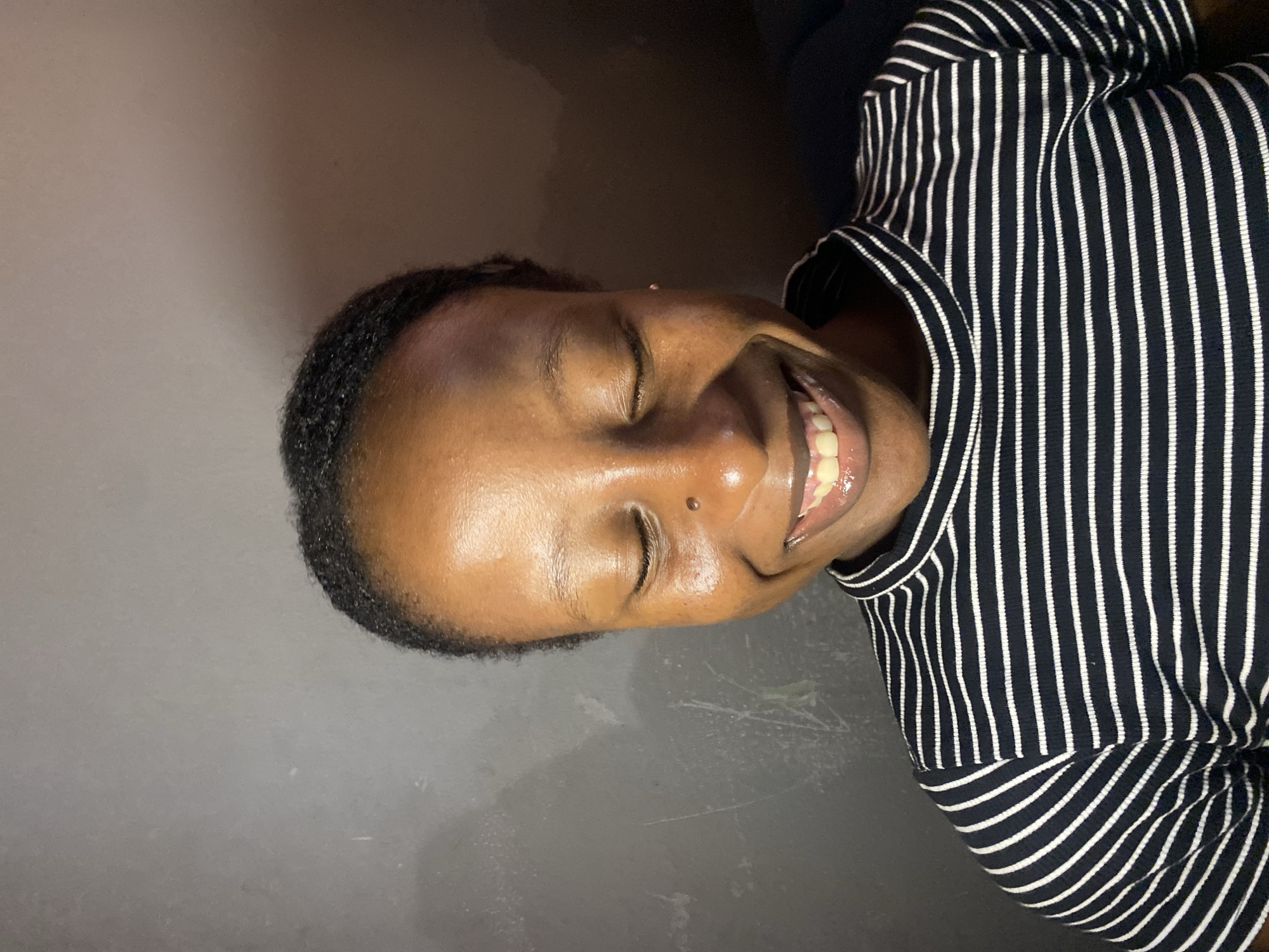 A medium shot photograph captures a young Black woman with her eyes closed, smiling broadly against a gray, textured background. She has short, dark hair pulled back from her face, and her skin appears dewy. A small piercing is visible on the bridge of her nose. She is wearing a black and white horizontally striped mock-neck top. The lighting is warm and appears to be coming from the right side of the frame, casting subtle shadows. The overall impression is one of joy and contentment.