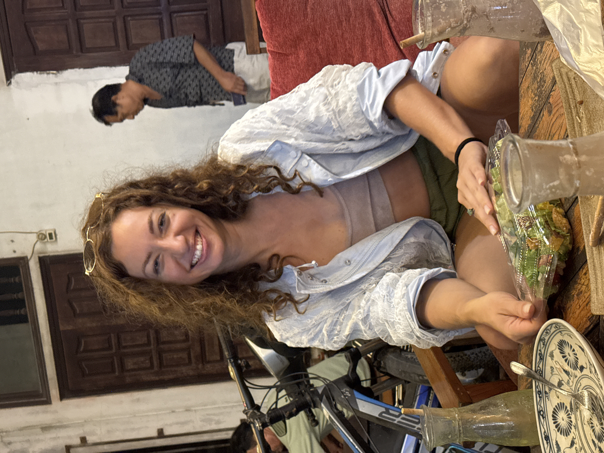A medium shot captures a smiling woman with curly brown hair, wearing glasses perched on her head, a light blue jacket over a light brown top, and green shorts. She is seated at a wooden table with a salad container and a fork in her hands. The background shows a rustic, dimly lit interior with wooden doors, a bicycle, and another person in the distance. The presence of what appear to be traditional Vietnamese architectural elements and the context of the surrounding environment suggest the location is likely within a historical or culturally significant area in Nam Hoa Lư Ward, Vietnam.