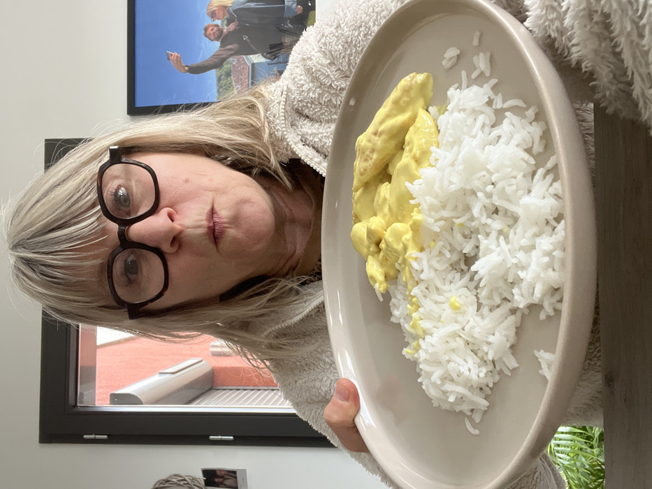 In Chambœuf, France, an adult female with blonde/light brown hair and dark-framed eyeglasses presents a meal indoors. She faces the camera directly, her mouth pursed and eyes wide. She wears a light-colored, textured jacket or sweater. Her left hand supports a round, light beige plate holding a serving of white rice next to pieces of a yellow, sauced dish, which partially covers the rice. To the subject's left, a window provides a view of a red building exterior with a grey downspout. Below the window, a woven decorative item and two small framed photographs are visible. To the subject's right, a large framed picture displays a man and a woman in an outdoor setting, taking a selfie. The man, bearded, holds a phone overhead while standing beside the woman. The interior walls are white, and a dark wooden surface is visible at the bottom of the frame.