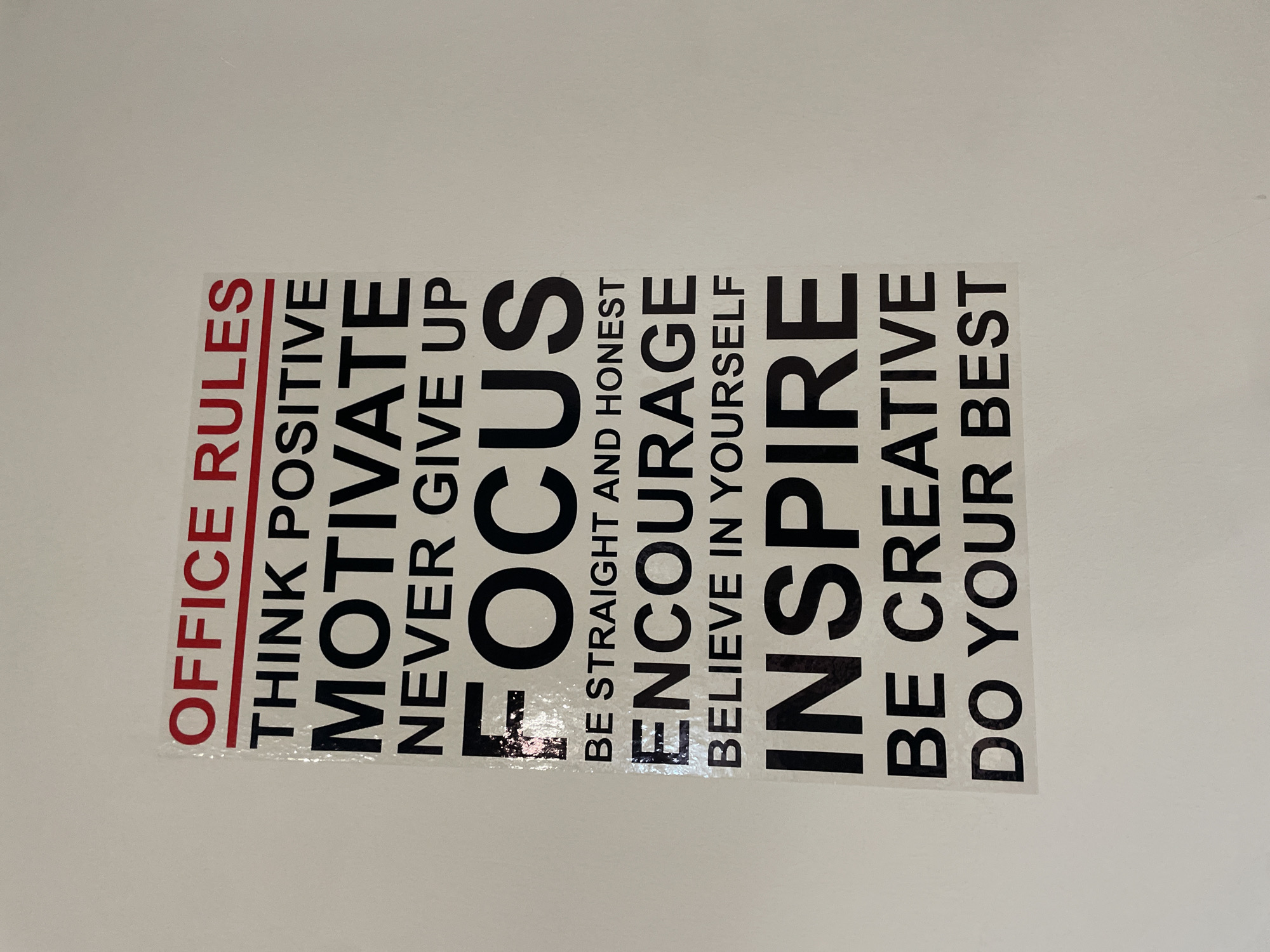 The image shows a motivational poster with "OFFICE RULES" in red text at the top, followed by a list of rules in black text on a white background. The rules are: THINK POSITIVE, MOTIVATE, NEVER GIVE UP, FOCUS, BE STRAIGHT AND HONEST, ENCOURAGE, BELIEVE IN YOURSELF, INSPIRE, BE CREATIVE, DO YOUR BEST. The poster is affixed to a plain, light-colored wall. The exact location within Tbilisi, Georgia, is not discernible from the image.