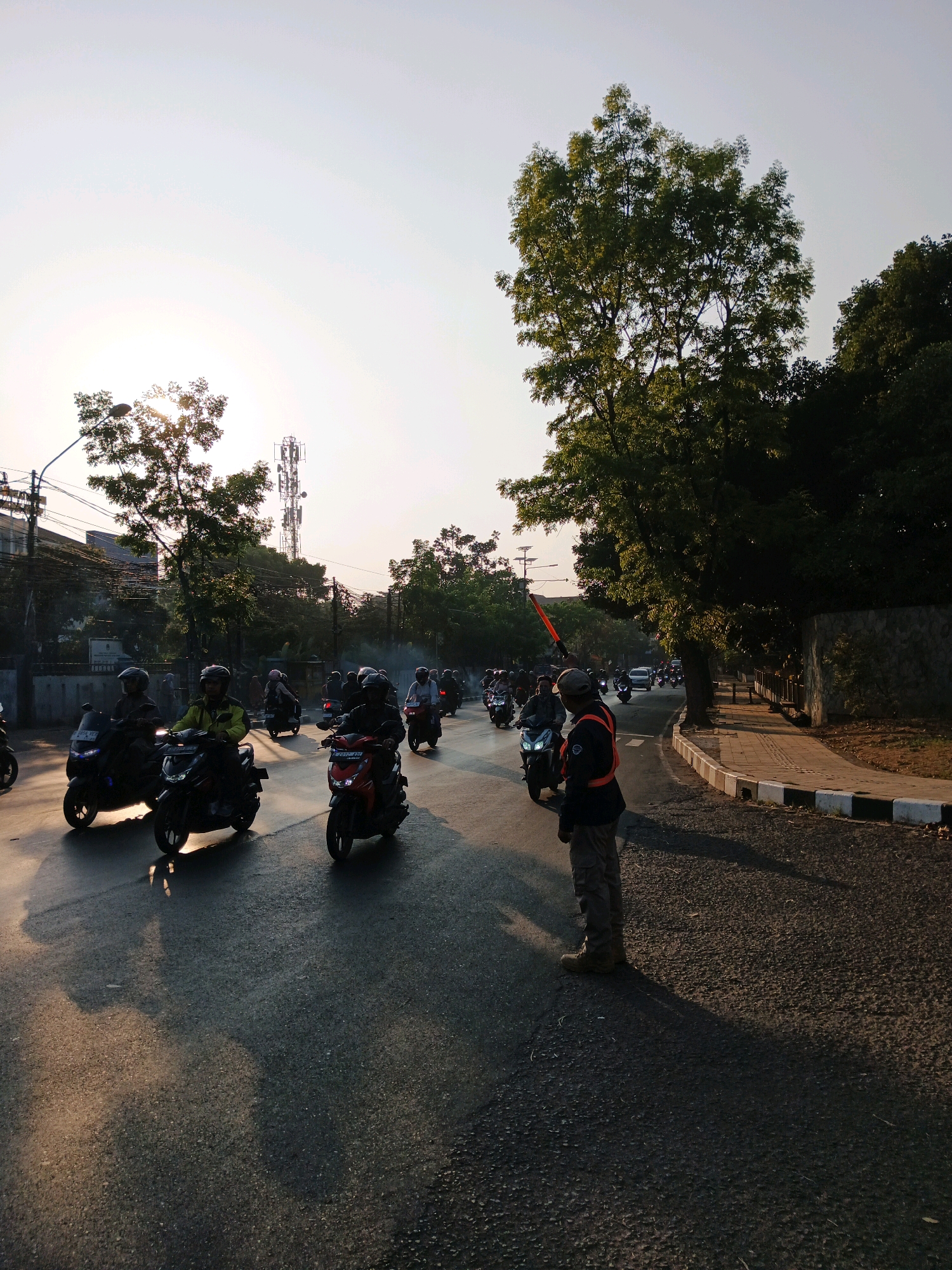 In a street scene in Bandung City, Indonesia, a traffic controller stands on the right side of the road, holding an orange signaling baton. He is wearing a high-visibility vest over a dark shirt and pants, with a cap and boots. The road is populated by a convoy of motorcycles with riders wearing helmets, suggesting a procession or heavy traffic. The sun is setting, casting long shadows and creating a warm, golden light that illuminates the scene from the left. Trees line the right side of the road, with a stone wall and sidewalk visible behind them. A cell tower is visible in the distance on the left, and power lines stretch across the sky. The atmosphere is that of a typical urban evening commute, with a touch of ceremonial order due to the traffic controller and the organized group of motorcycles.